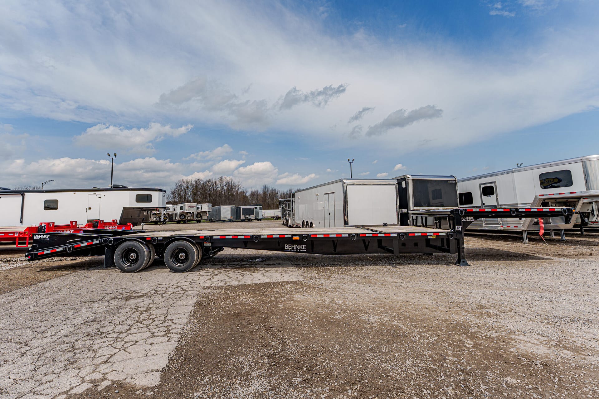 Long black flatbed trailer parked in a gravel lot with multiple white trailers in the background under a blue sky.