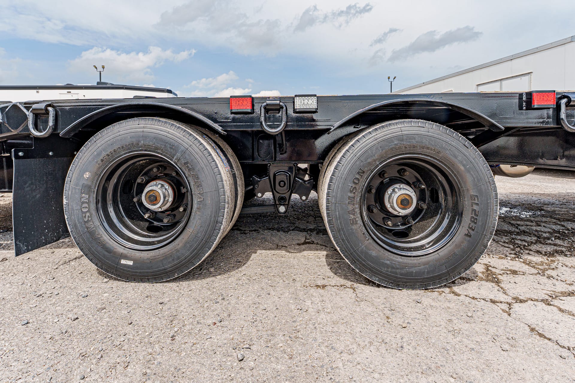 Dual rear trailer wheels on a black flatbed with mud flaps, parked on a gravel lot.