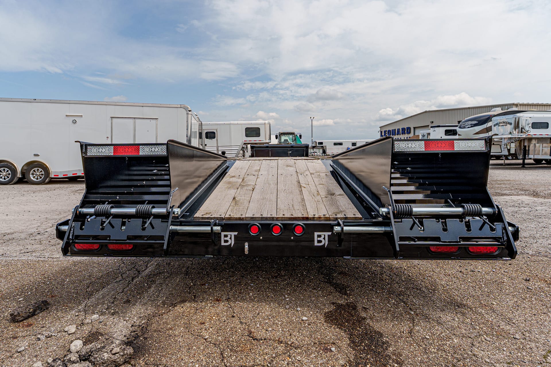 Wide view of a black flatbed trailer with wood loading ramp in a truck yard, ready for loading vehicles and equipment.