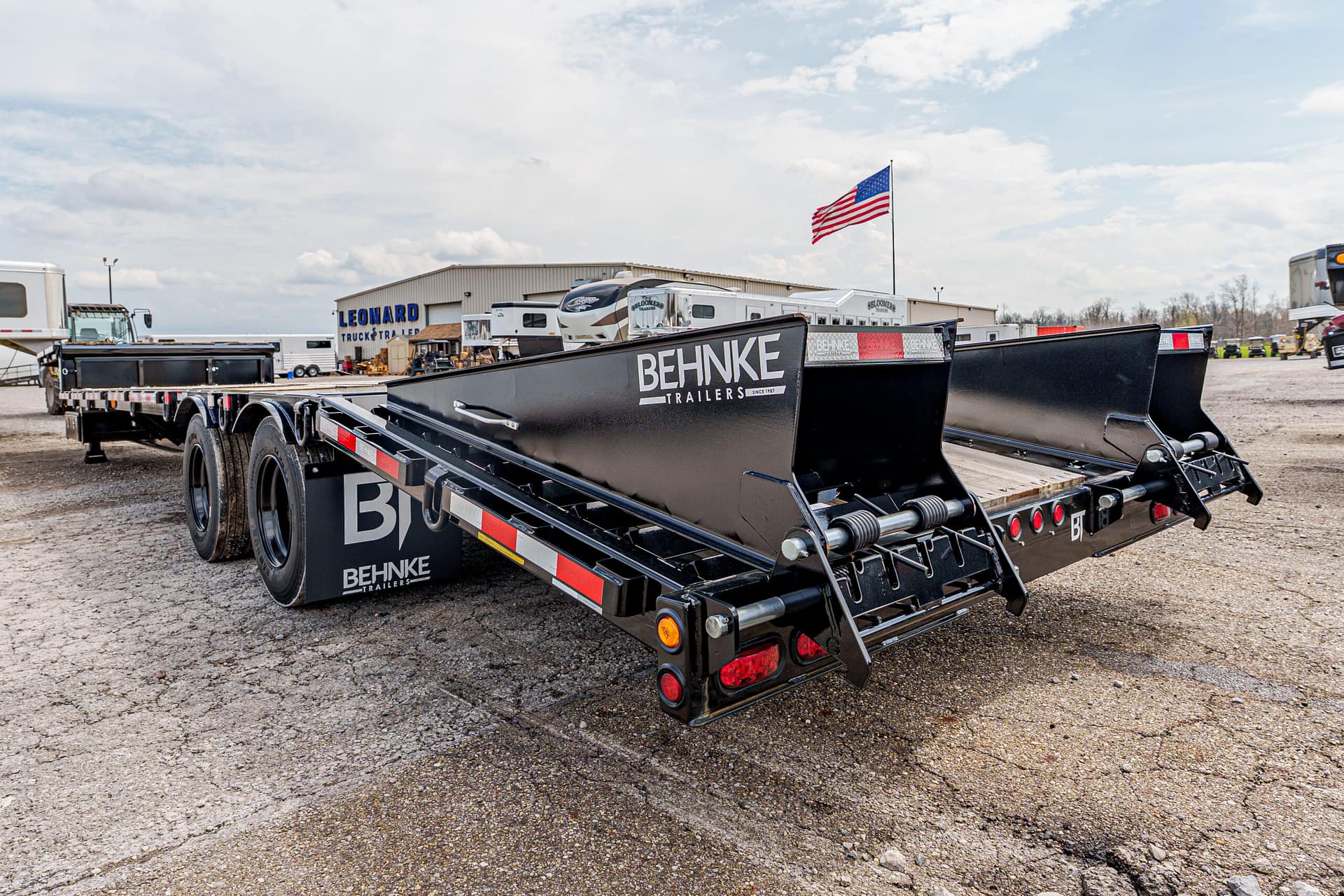 Black Behnke flatbed trailer with loading ramps in a dealership lot, American flag in the background.