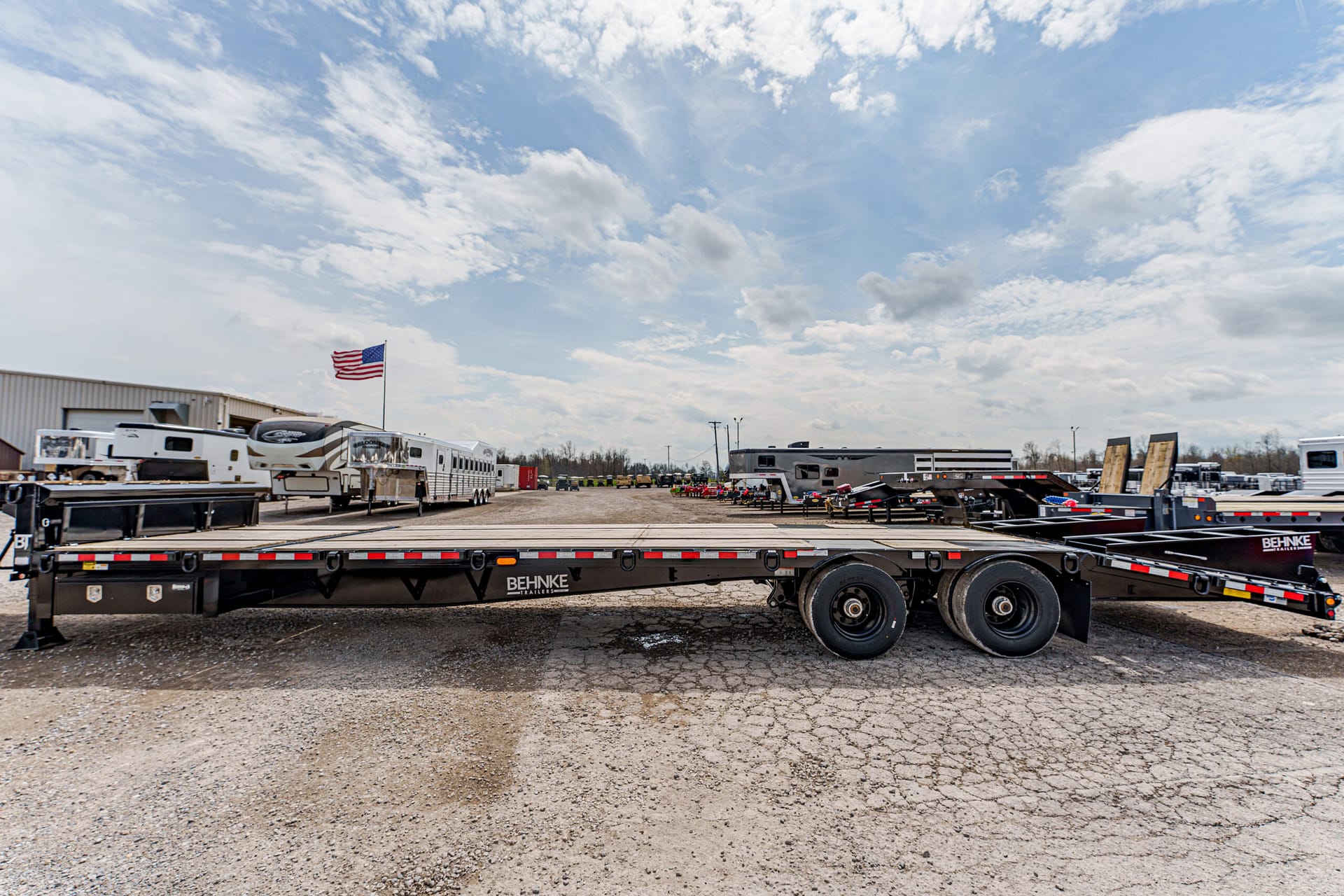 Long black flatbed trailer in a truck yard with an American flag and rows of RVs and trailers in the background.