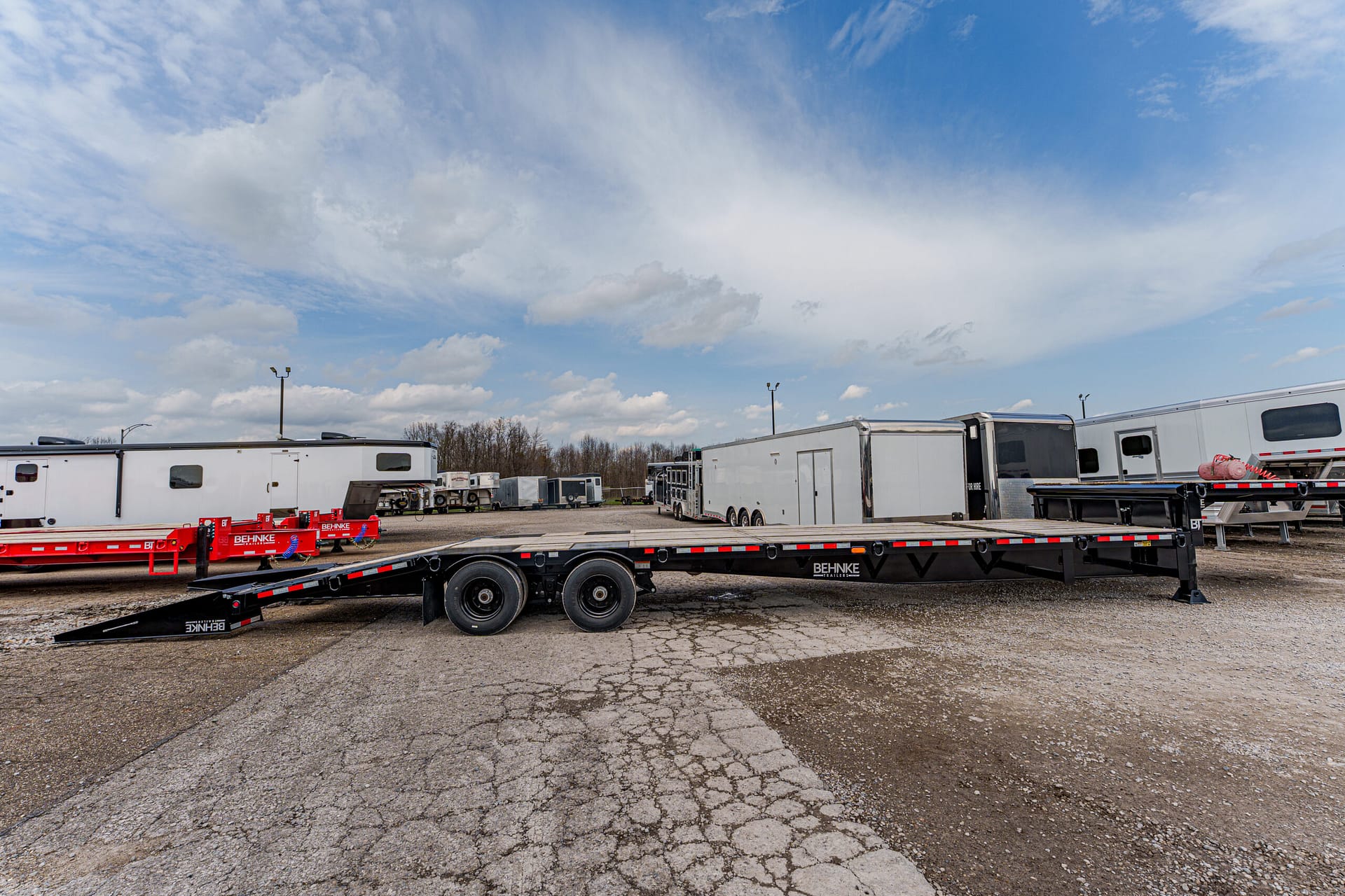 Long flatbed trailer in a truck yard with other trailers and storage buildings in the background, under a blue sky with scattered clouds.