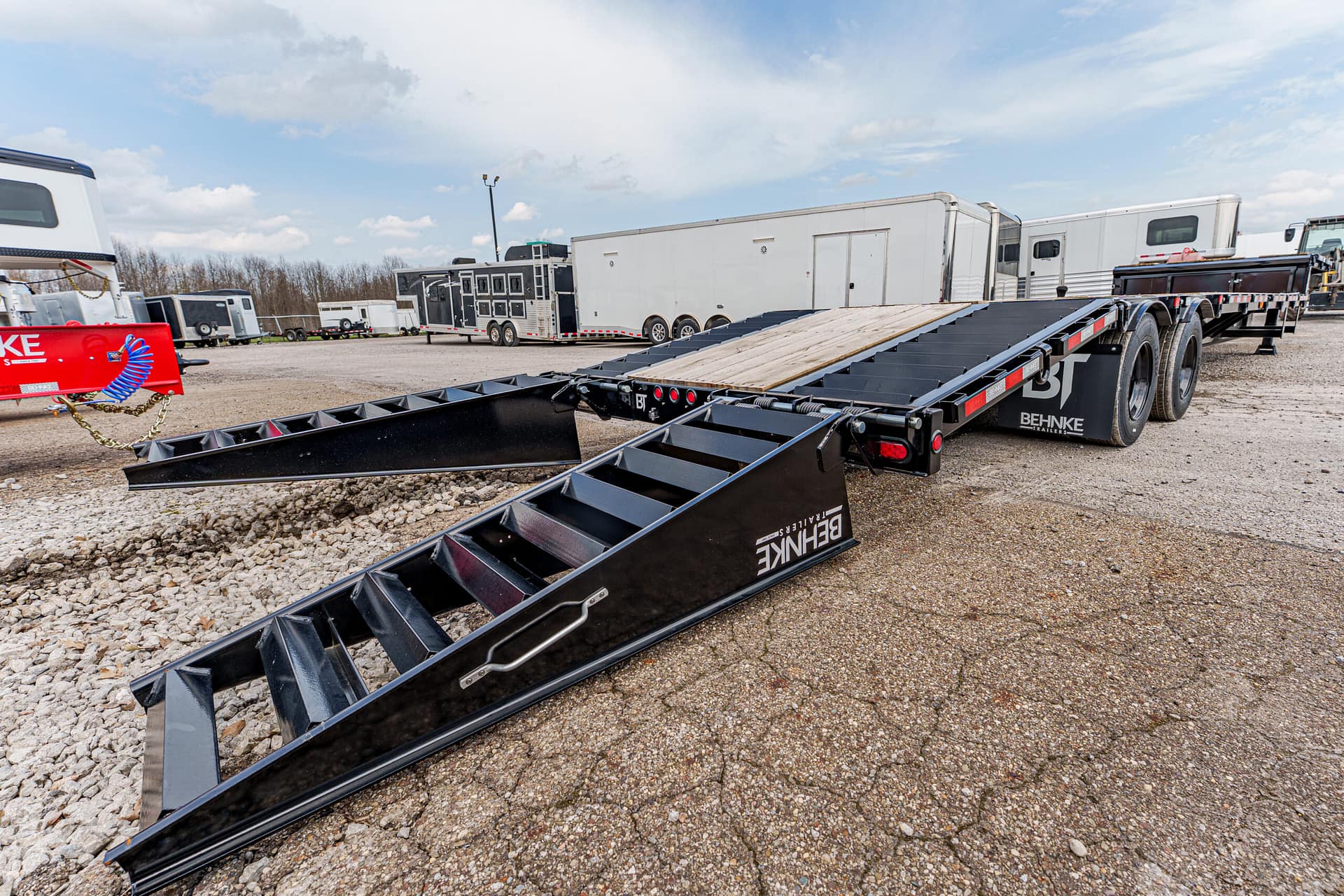 Black Behnke lowboy trailer with dual loading ramps extended, parked on a gravel lot with other trailers in the background, ready for loading.
