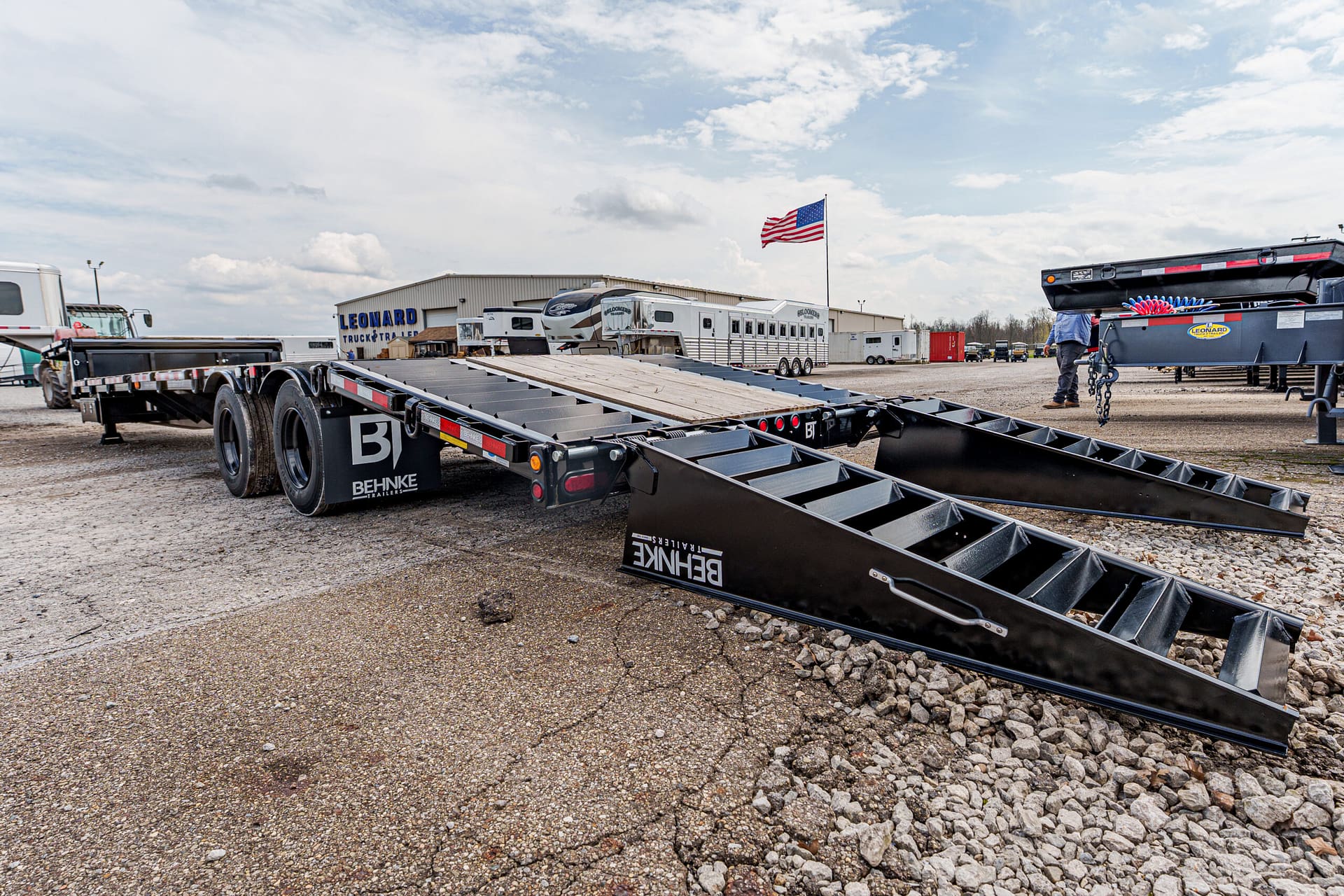 Flatbed trailer with metal loading ramps in a truck yard, Behnke Trailers branding visible, American flag flying in the background