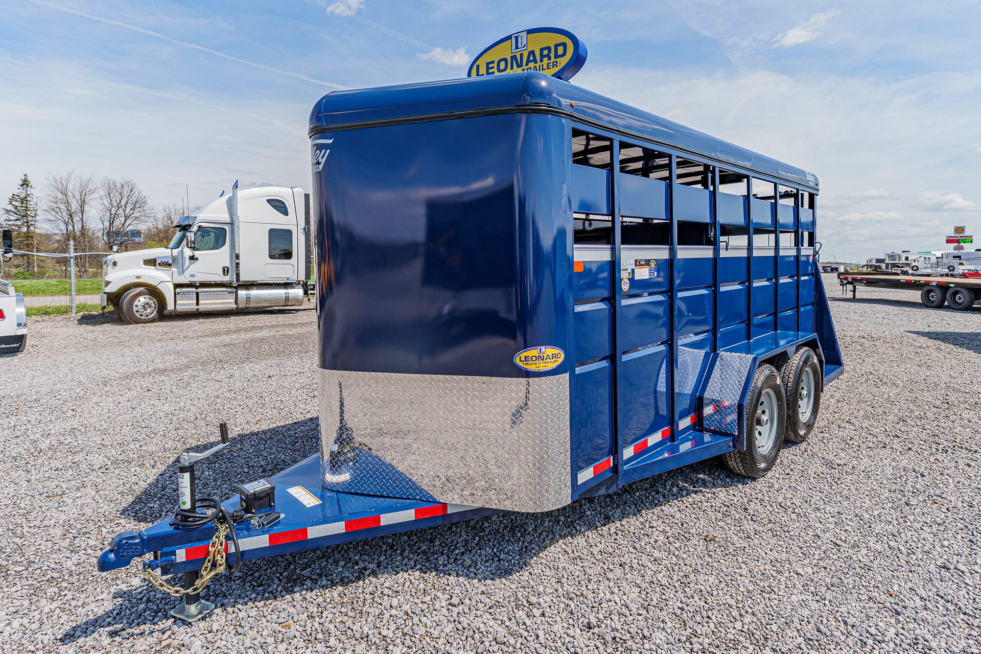 Blue livestock trailer with diamond-plate front and dual-axle on a gravel lot, hitch at the front ready to tow with a white truck nearby in the background