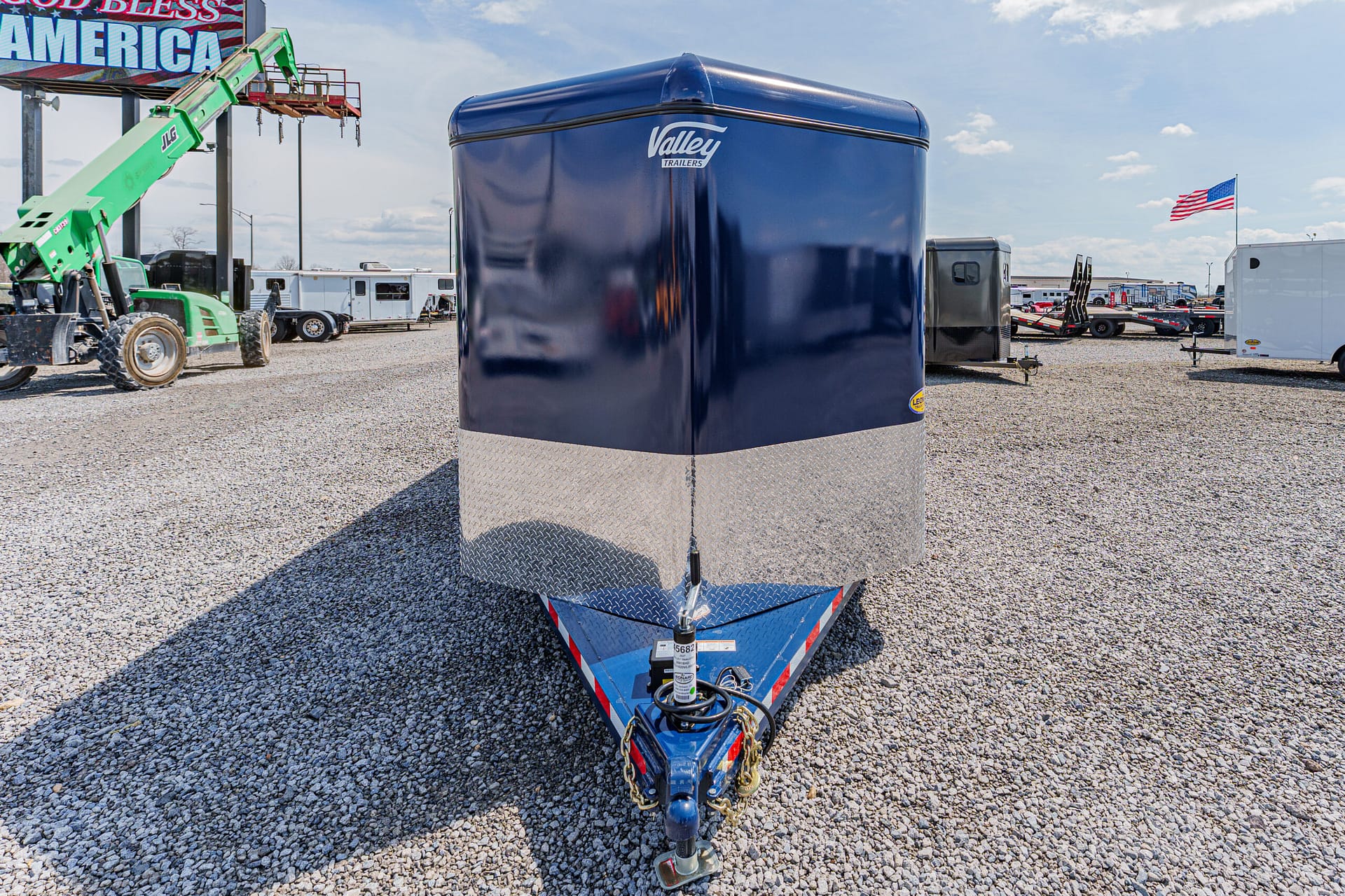 Blue enclosed cargo trailer with diamond-plate lower front sits on a gravel lot, hitch connected, with other trailers and equipment in the background.