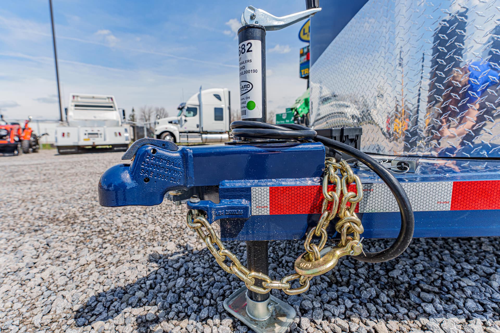 Close-up of a blue trailer hitch with a safety chain and lock on a gravel lot. Infers trailer is secured and awaiting hitching.