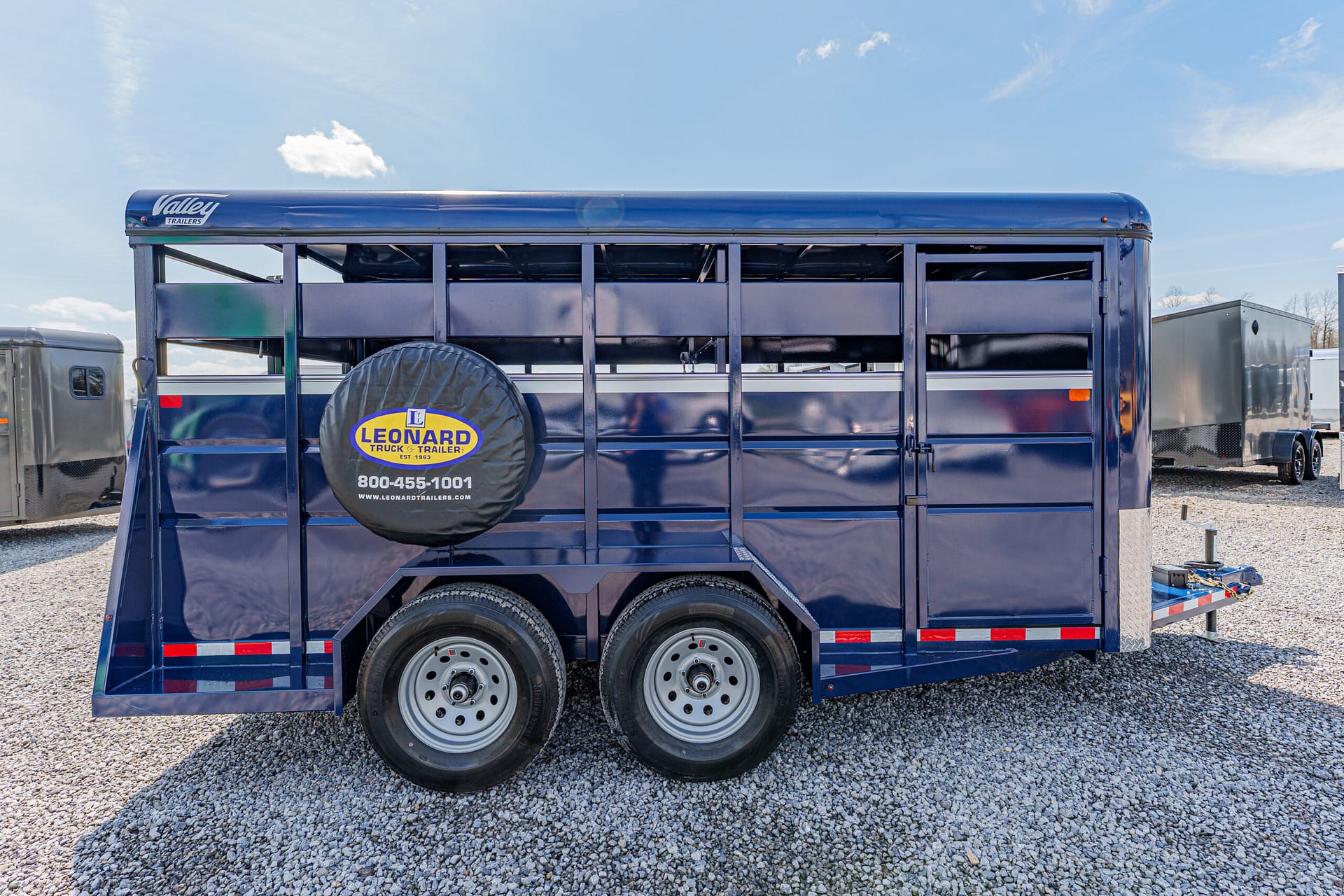 Blue horse trailer with a spare tire cover reading Leonard Trailer, parked on a gravel lot (dual axles, dark blue finish).