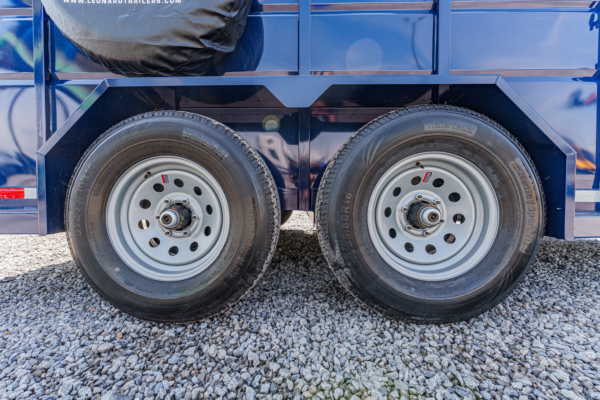 Dual trailer wheels on a blue trailer parked on a gravel surface with a spare tire mounted above the wheels