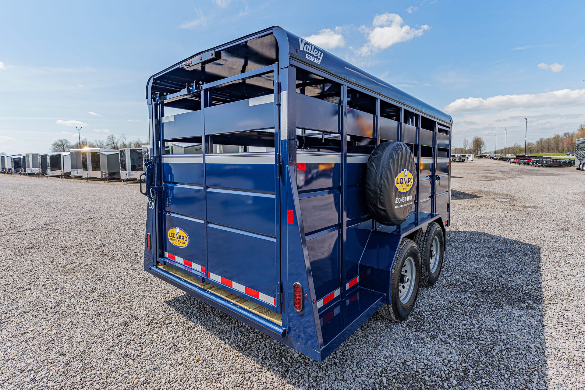 Rear of a blue two-axle livestock trailer on a gravel lot, spare tire mounted at the back and a ramp door visible.