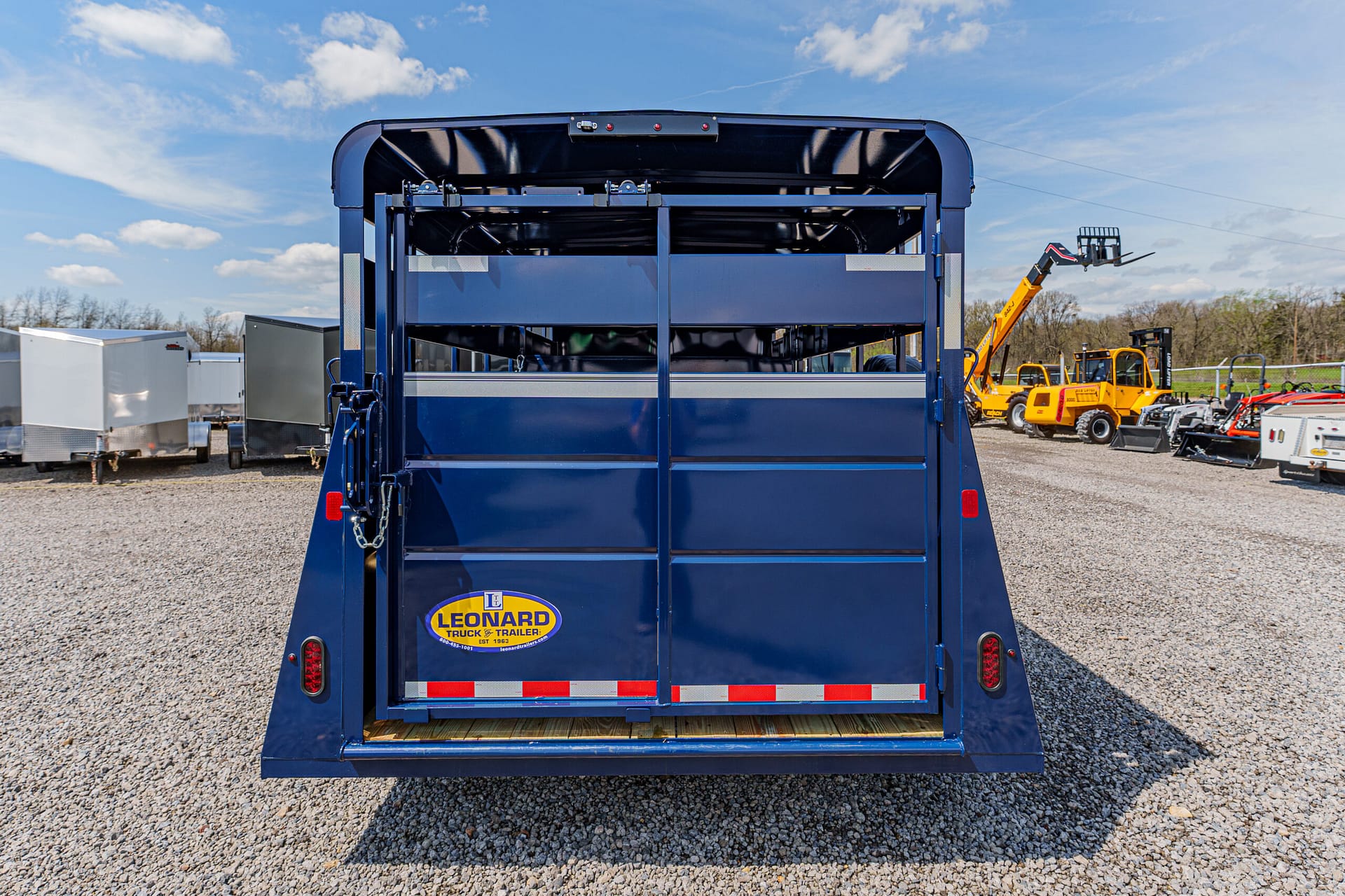 Rear of a blue Leonard truck trailer parked on a gravel lot, with other trailers and yellow machinery in the background.