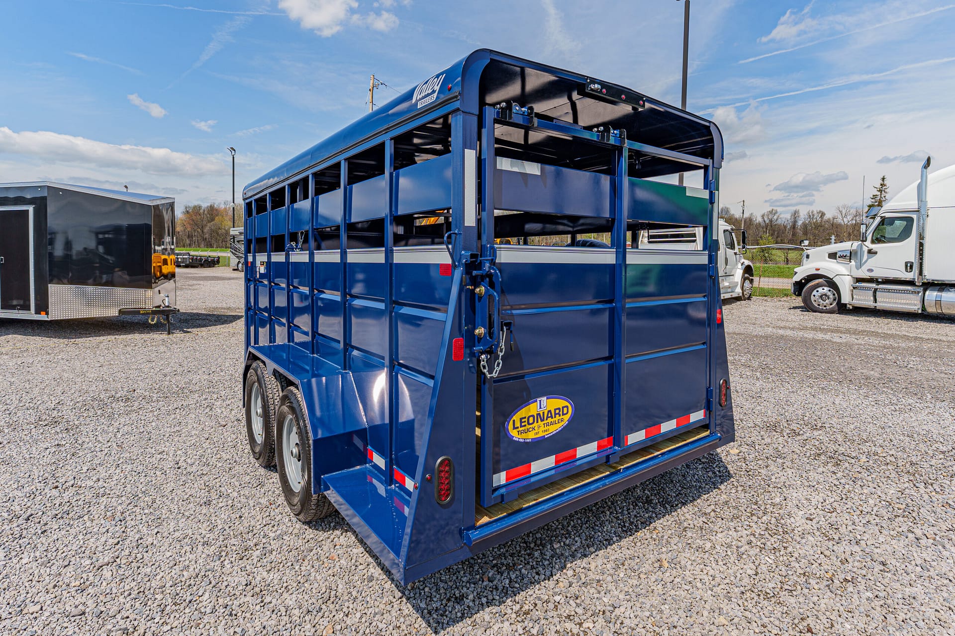 Blue livestock trailer with dual axles and rear gate in a gravel lot; Leonard Truck & Trailer logo visible on back panel