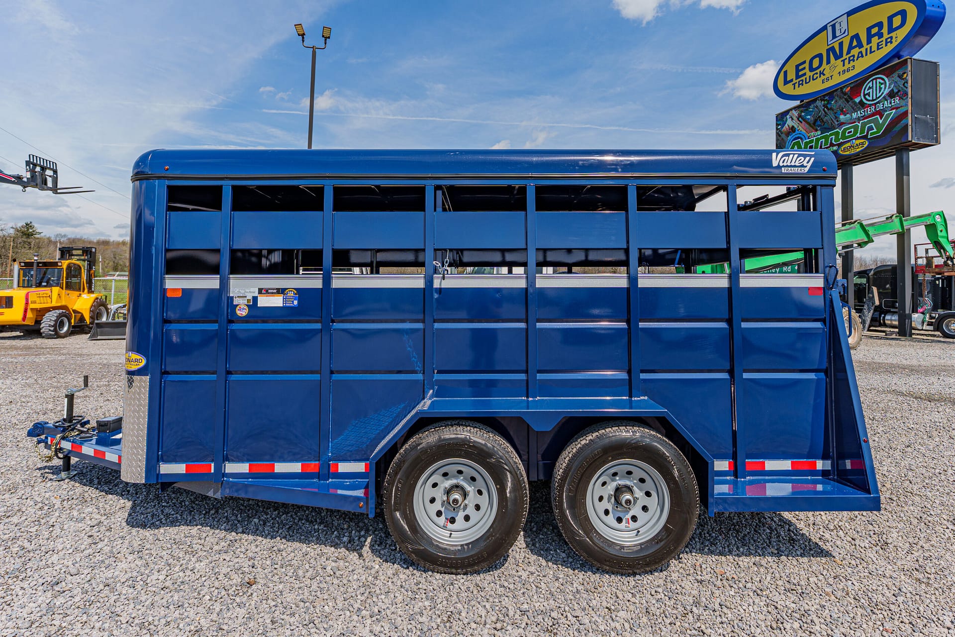 Blue tandem-axle livestock trailer parked on a gravel lot with farm equipment and a sign in the background, used for transporting animals