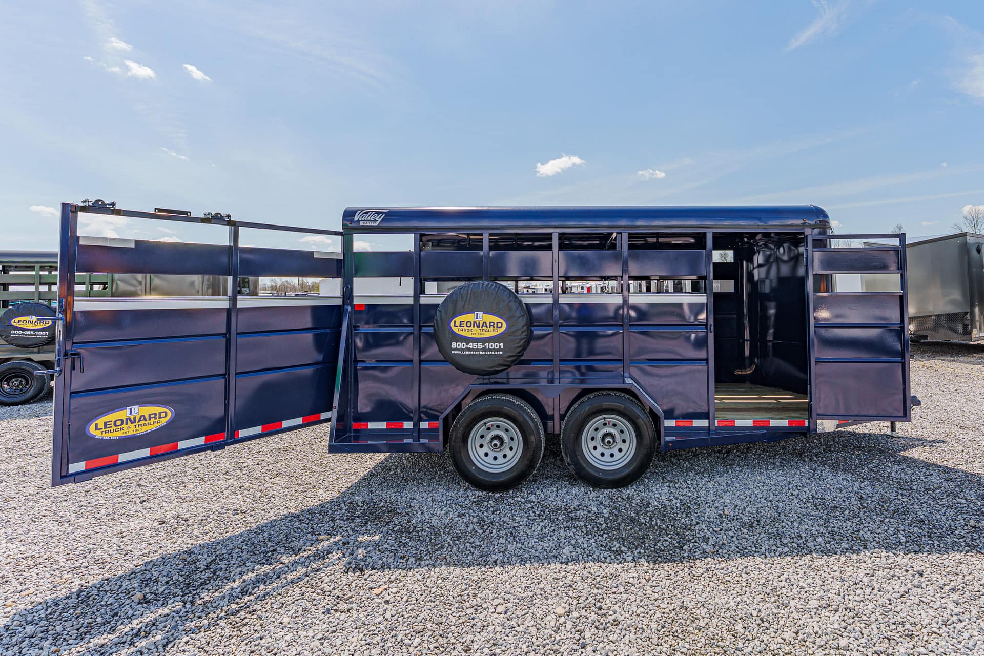 Blue double-axle livestock trailer with side doors open and spare tire mounted on the side, on a gravel lot under a clear sky, Leonard branding visible.