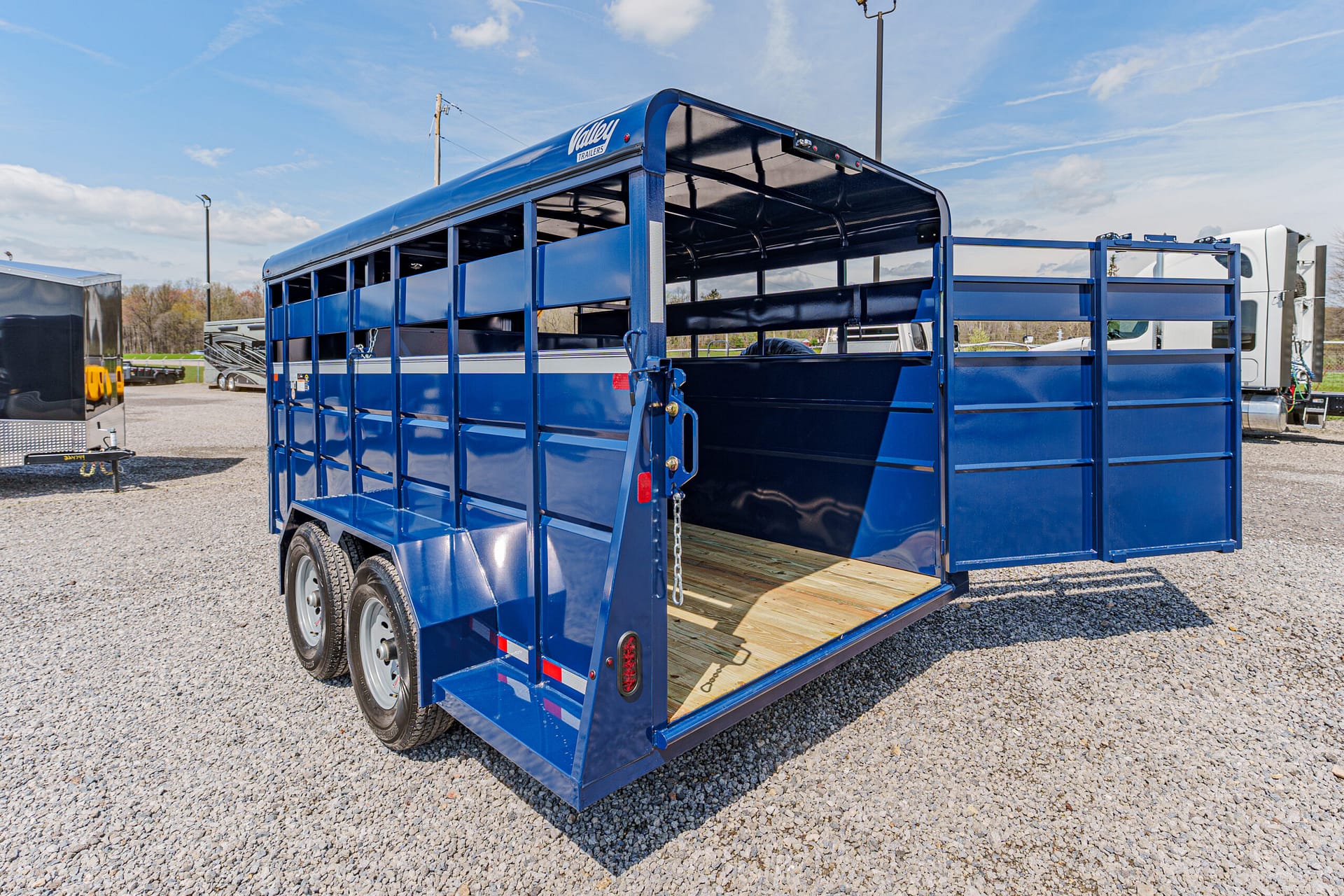 Blue and dark-blue livestock trailer with dual axles and open rear gate on a gravel lot; wooden floor visible inside.