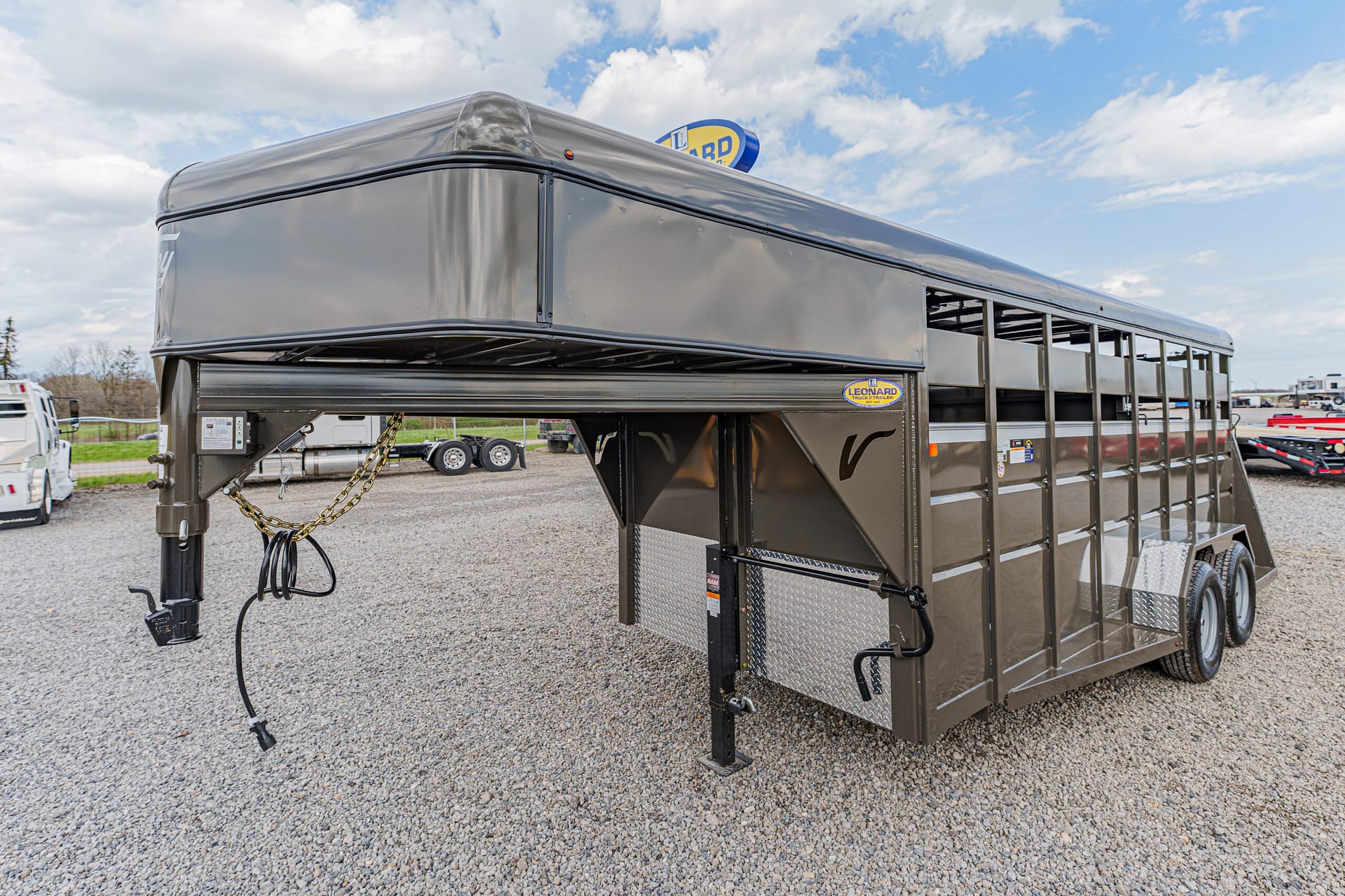 Dark gray gooseneck livestock trailer with dual-axle setup, parked on a gravel lot with a chrome diamond-plate kick plate and safety hitch visible in the foreground.