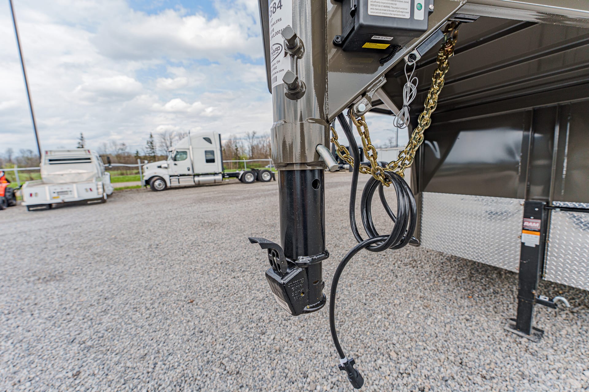 Gooseneck trailer hitch with attached safety chains and electrical wires hanging from the hitch in a truck yard.