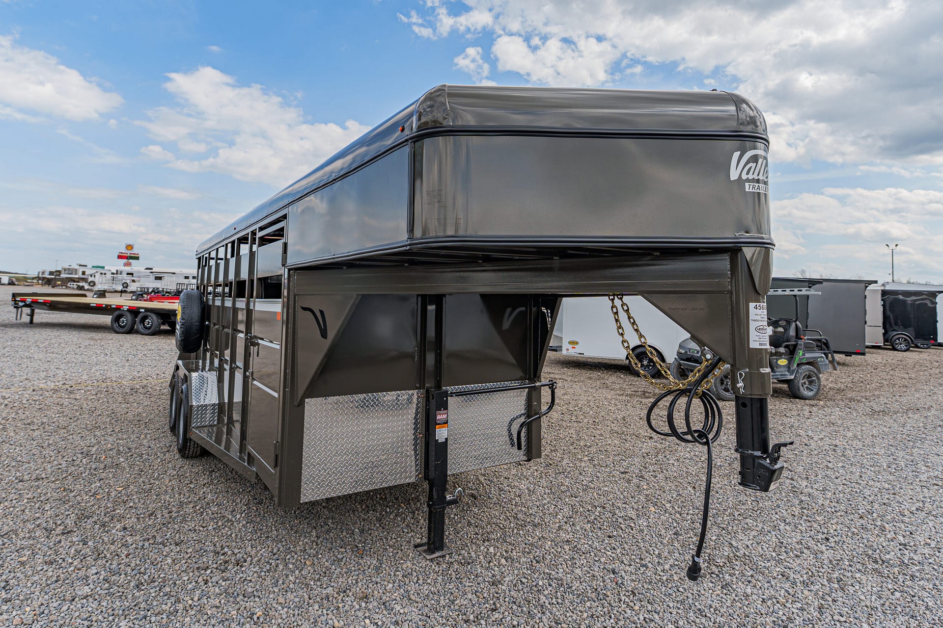 Gray gooseneck horse trailer with dual axles and diamond-plate storage on a gravel lot; other trailers in the background.
