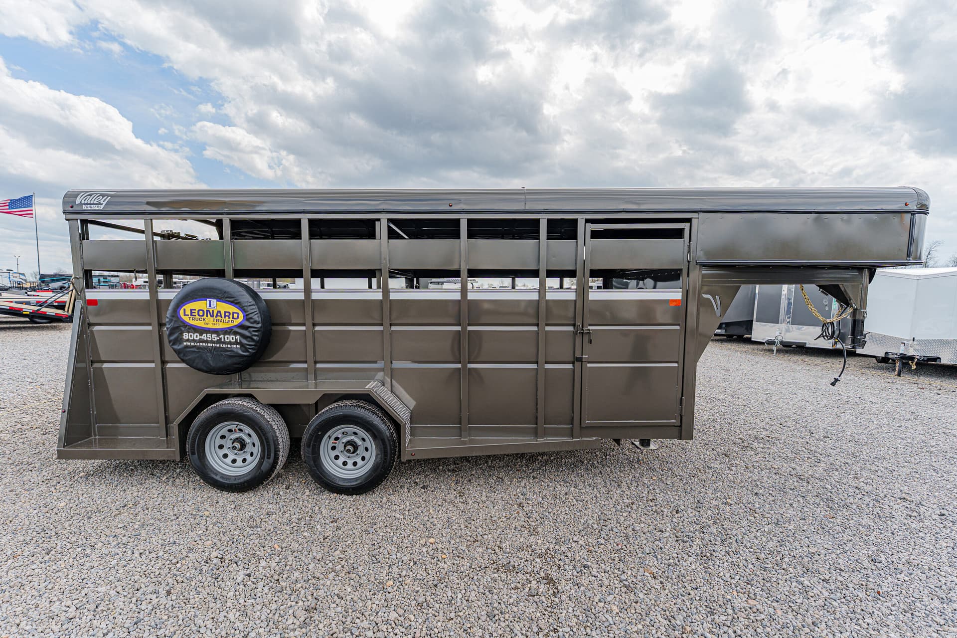 Gooseneck livestock trailer with dual axles and spare tire mounted on the side, parked on a gravel lot under a cloudy sky, barn-style vents along the top edge.