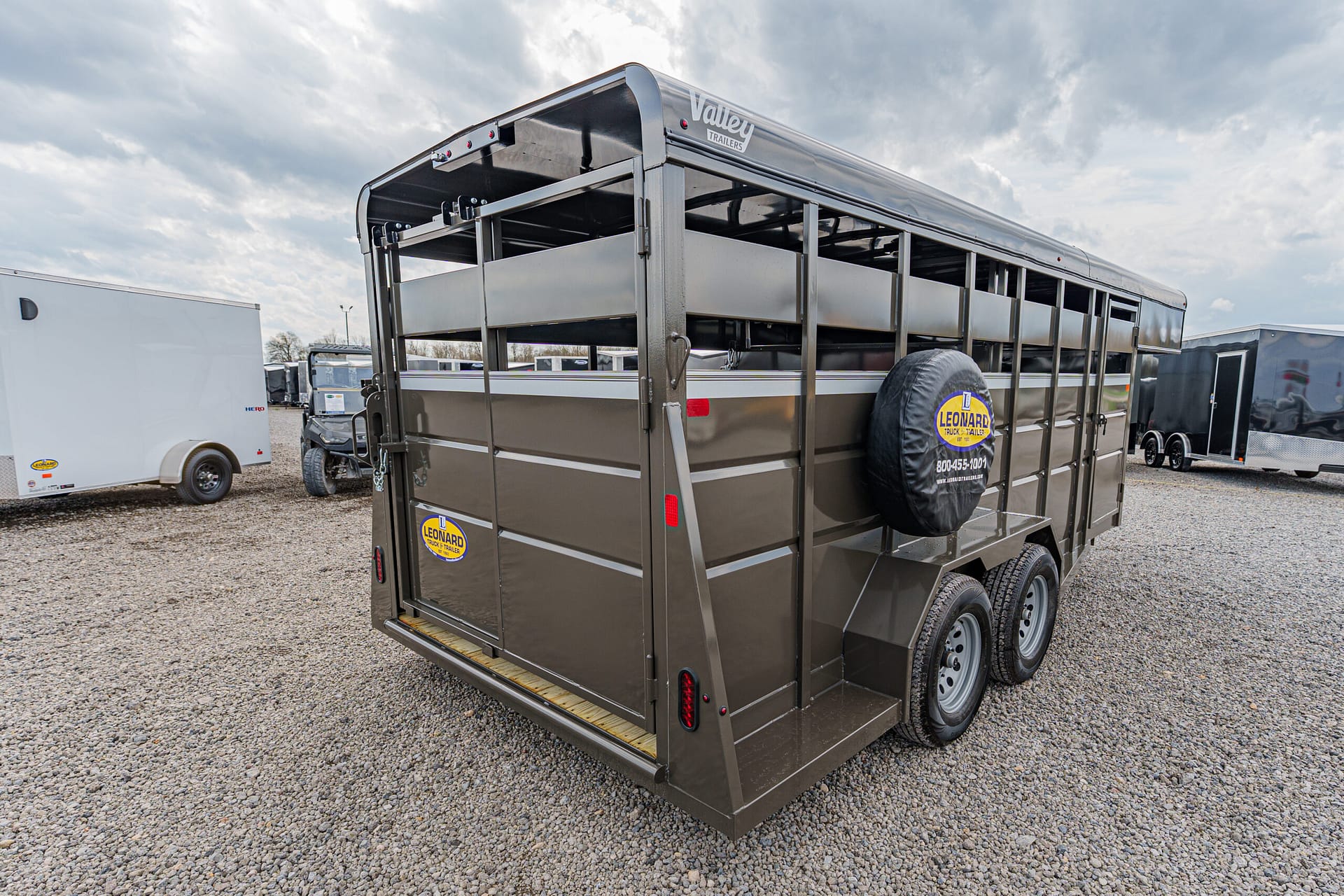 Brown livestock trailer with dual axles and a spare tire mounted on the side, parked on a gravel lot under cloudy sky (Valley Trailers logo visible).