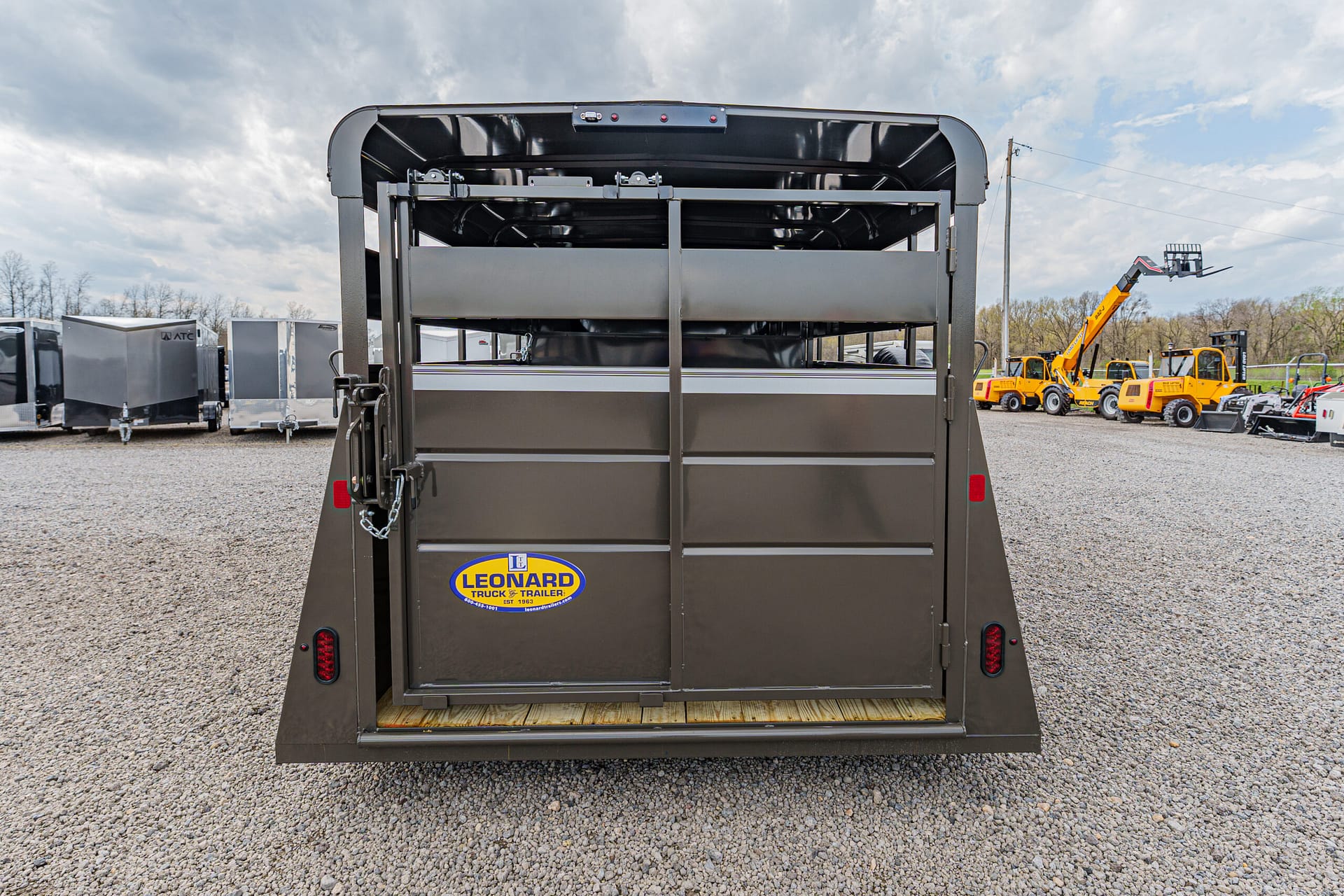 Rear view of a dark brown livestock/trailer with a vertical divider, taillights, and a Leonard Truck & Trailer sticker in a lot of equipment trailers behind it