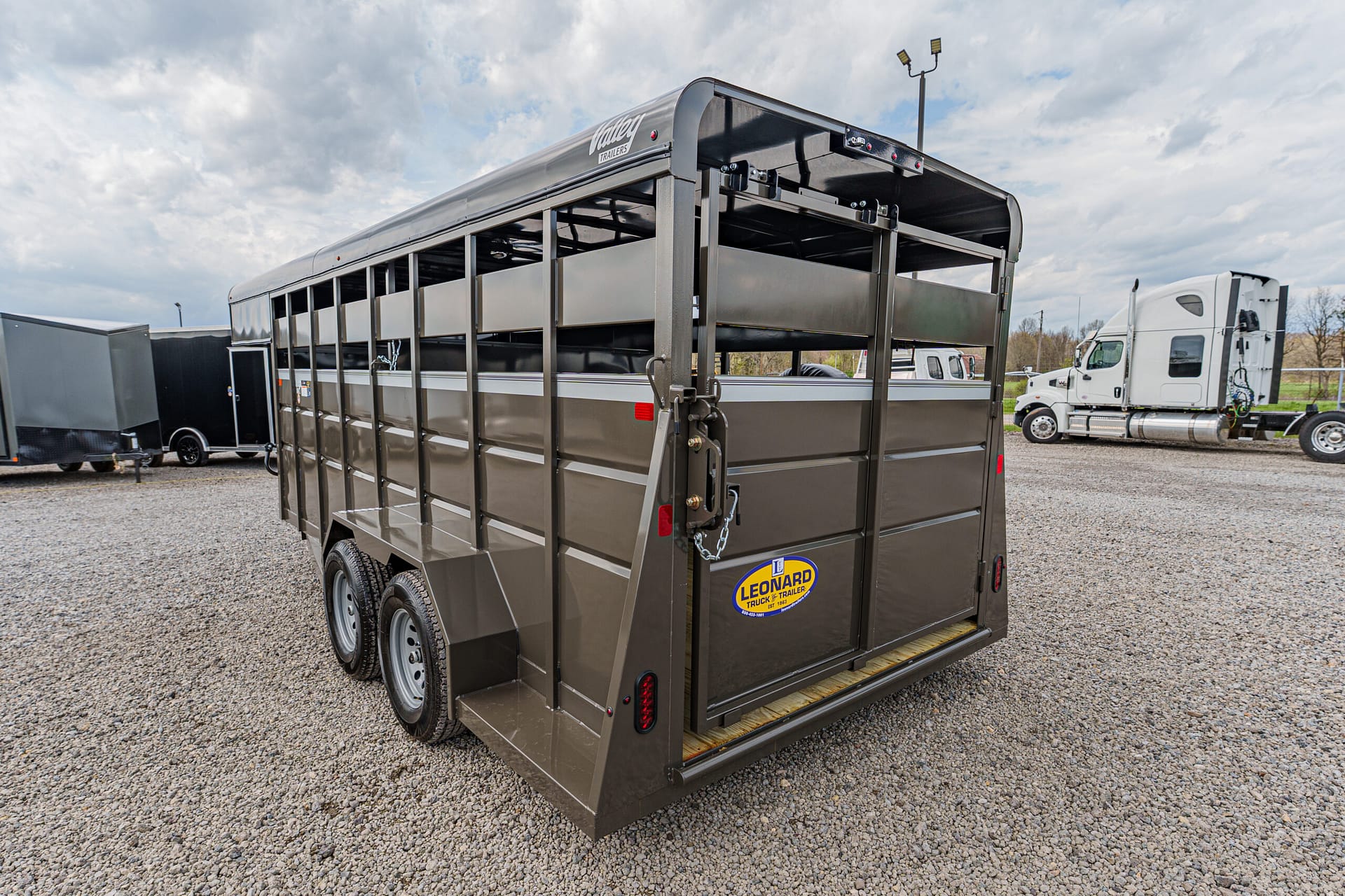 Two-axle brown steel livestock trailer with slatted sides and a rear ramp, parked on a gravel lot (Leonard trailer logo visible).