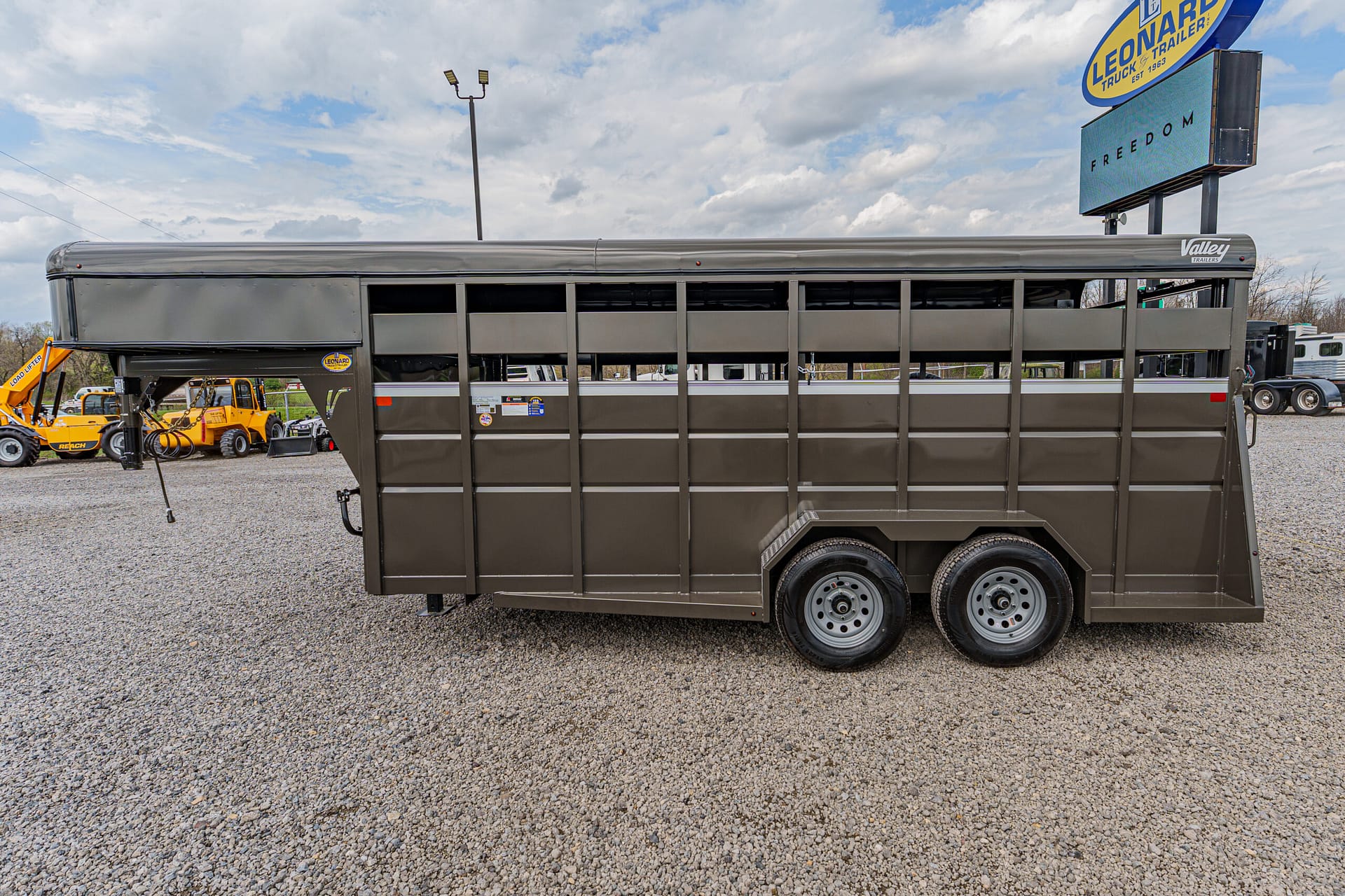 Gray dual-axle livestock trailer on a gravel lot with open side panels and equipment in the background.