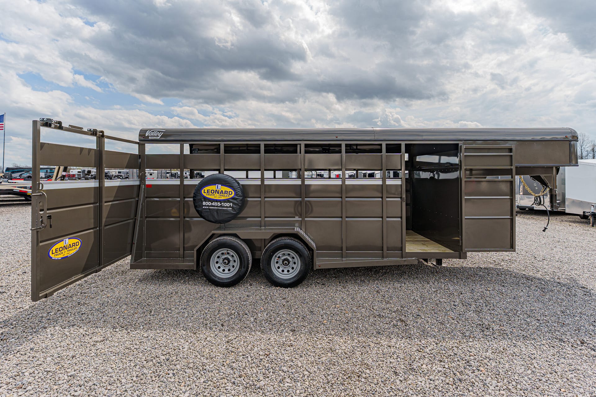 Brown dual-axle livestock trailer with open side door, showing a spare tire cover that reads Leonard Trailer, parked on gravel lot
