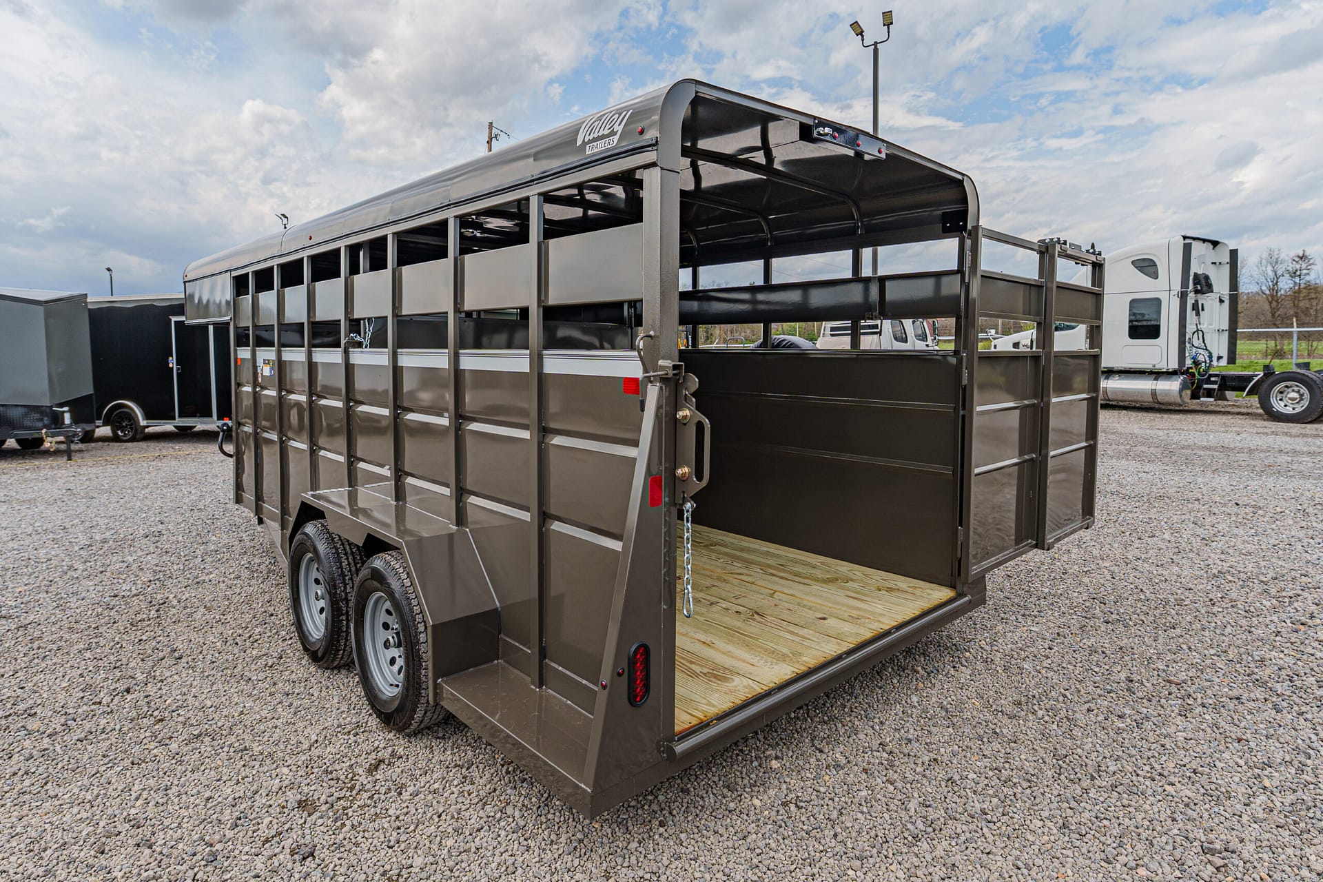Two‑axle brown livestock trailer with its rear ramp lowered onto a gravel lot, wooden floor visible inside.