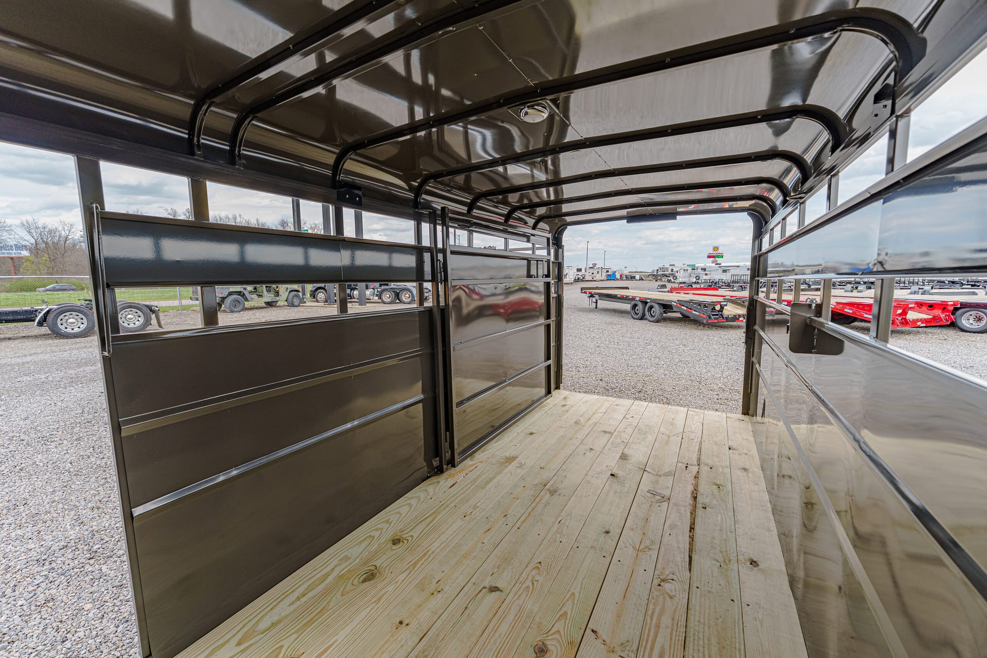 Interior of a black metal utility trailer with a wooden plank floor, open sides, and overhead rails, facing a gravel lot with other trailers outside