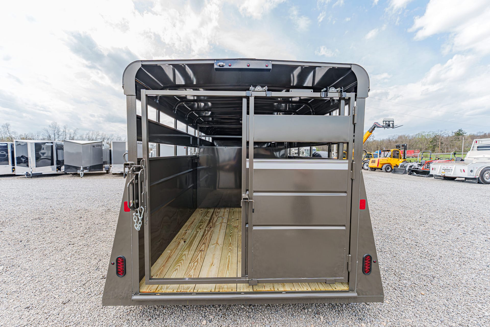 Back of a dark gray livestock trailer with the rear doors open, showing a wooden floor and divider inside on a gravel lot with other trailers nearby.
