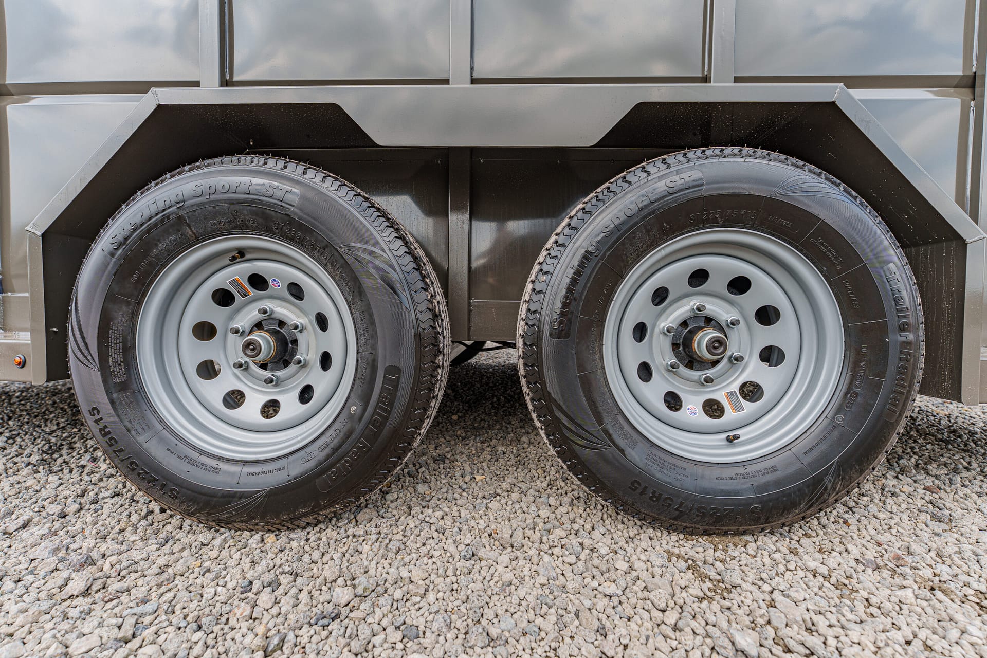Dual trailer wheels on a gravel surface under a metal trailer frame, showing rims and tires side by side.