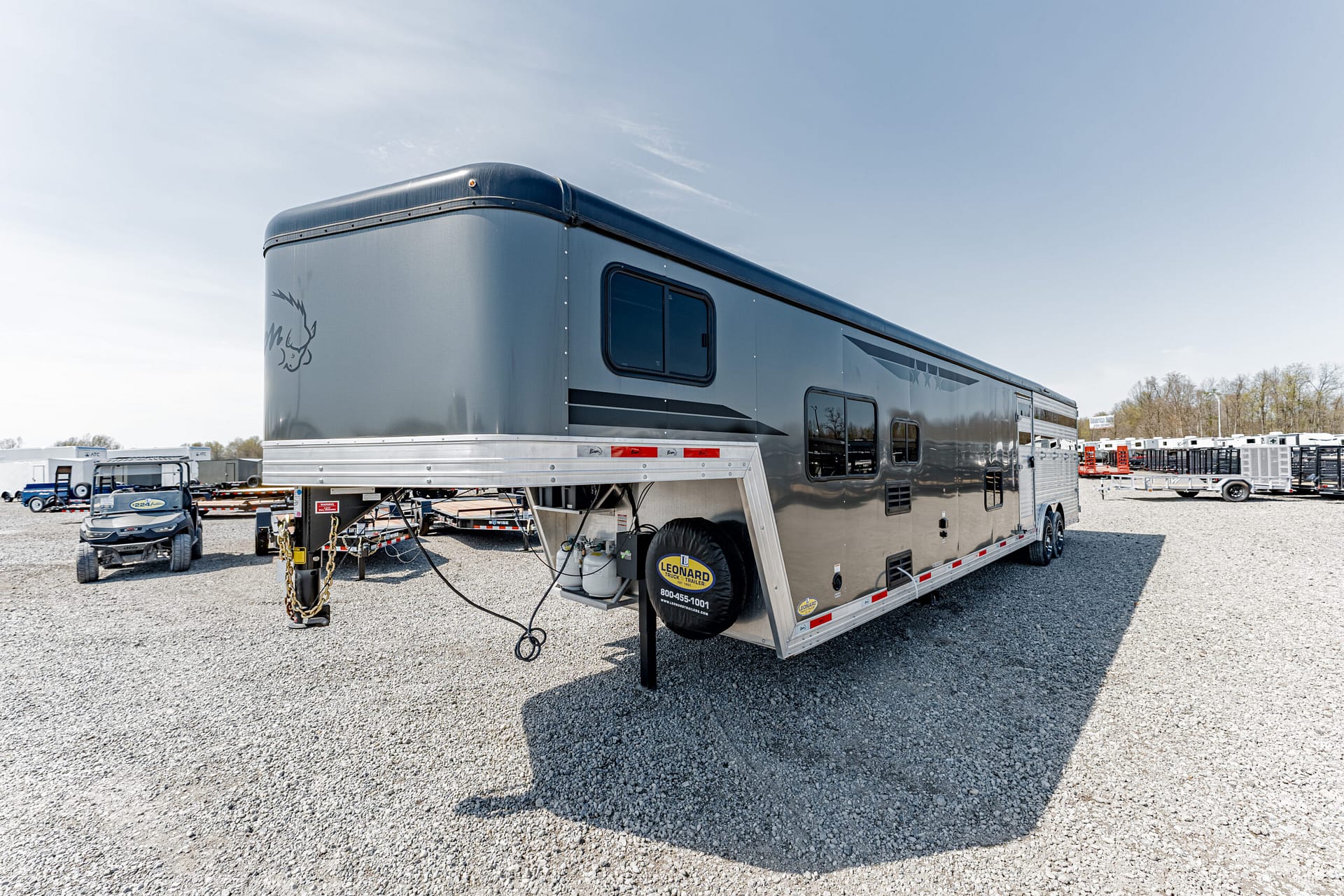 Front corner of a large gray fifth-wheel trailer parked on a gravel lot, with other trailers in the background.