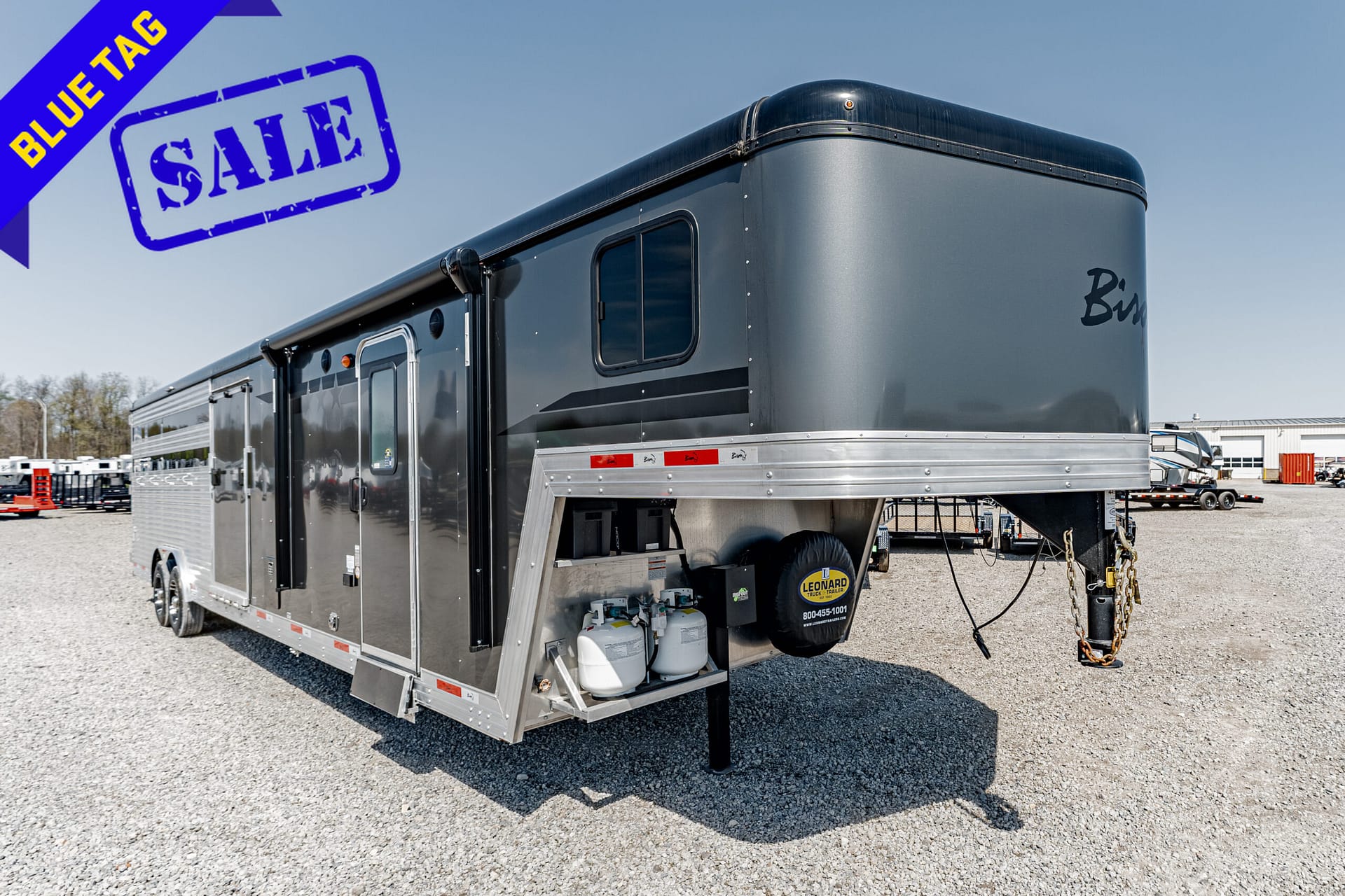 Gray gooseneck trailer on a gravel lot with a large blue 'Blue Tag' sale banner in the top-left corner