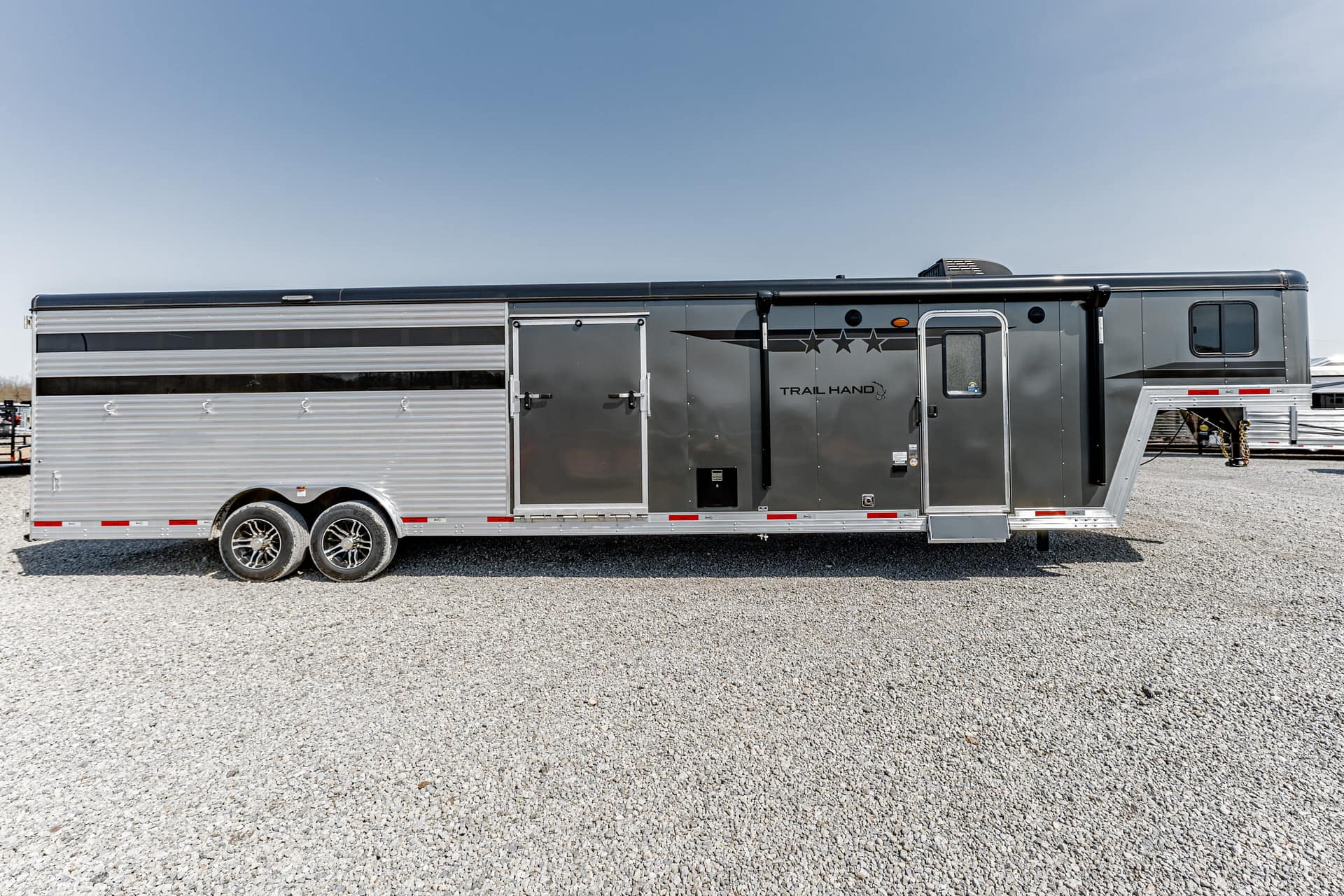 Long silver horse trailer with three axles, side doors, and 'Trail Hand' branding parked on a gravel lot under a clear blue sky.
