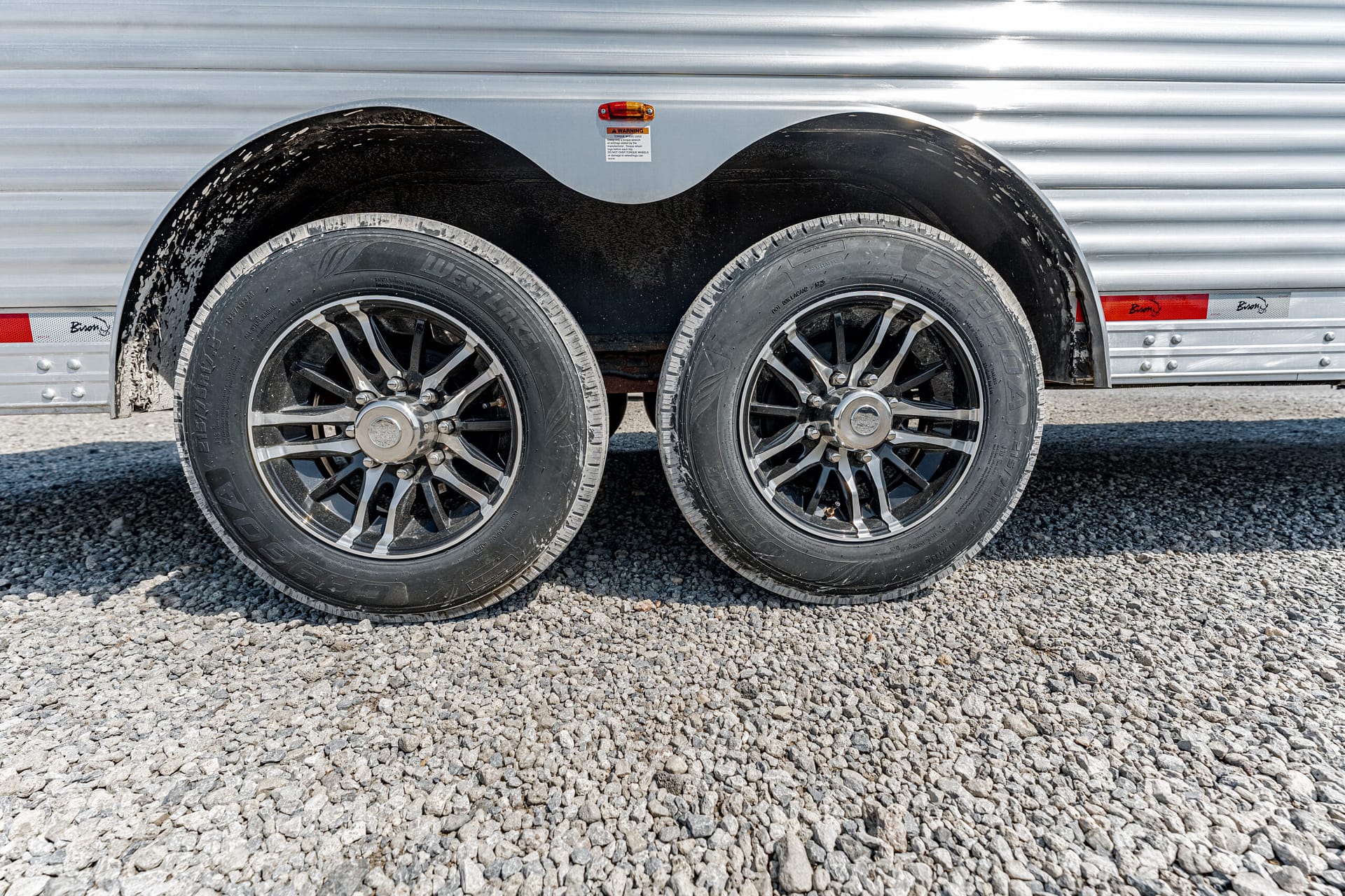 Dual trailer wheels with tires mounted on a gravel surface, under a silver metal trailer with a curved fender