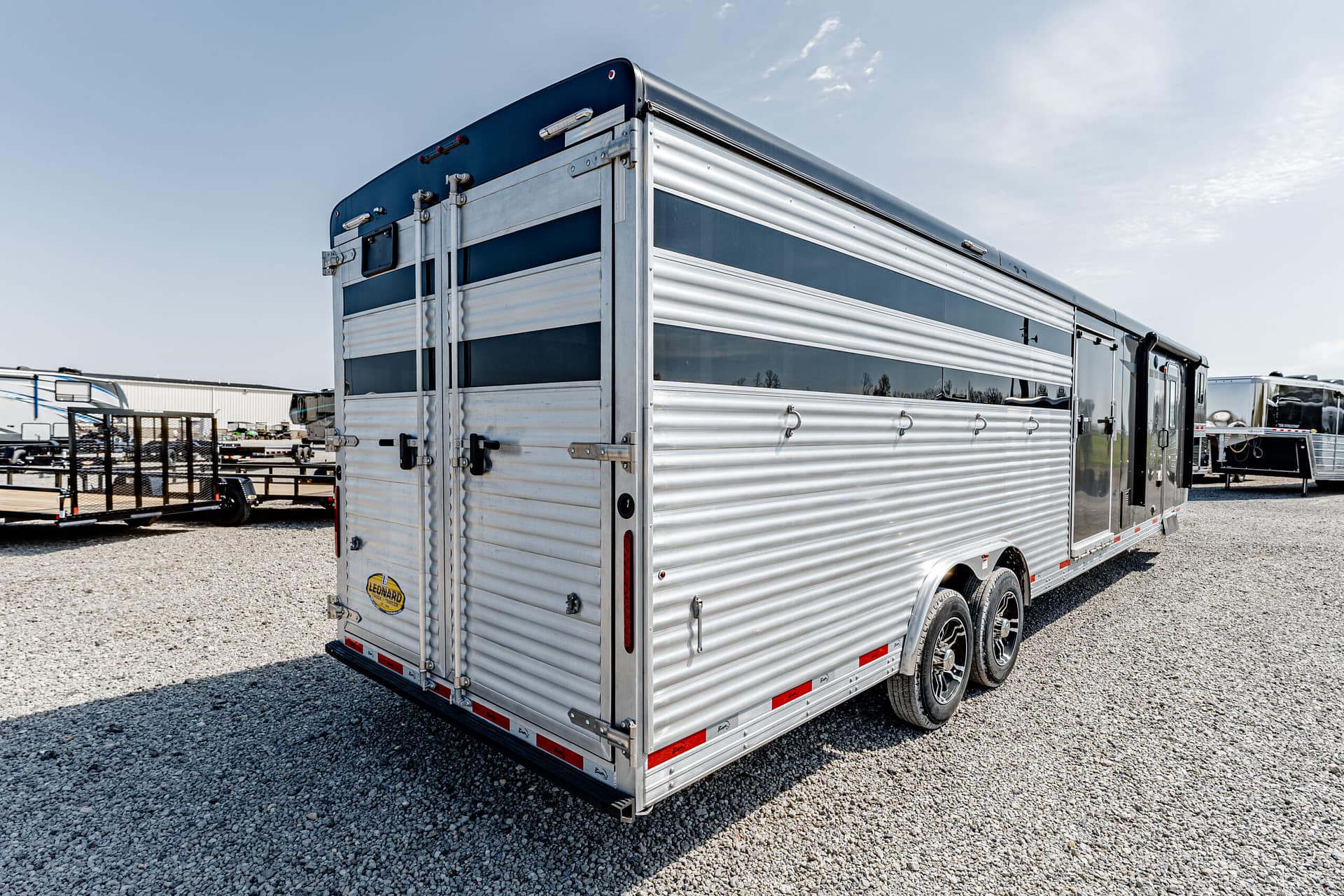 Aluminum travel trailer with ribbed siding and multiple windows, parked on a gravel lot with other trailers in the background in daylight.