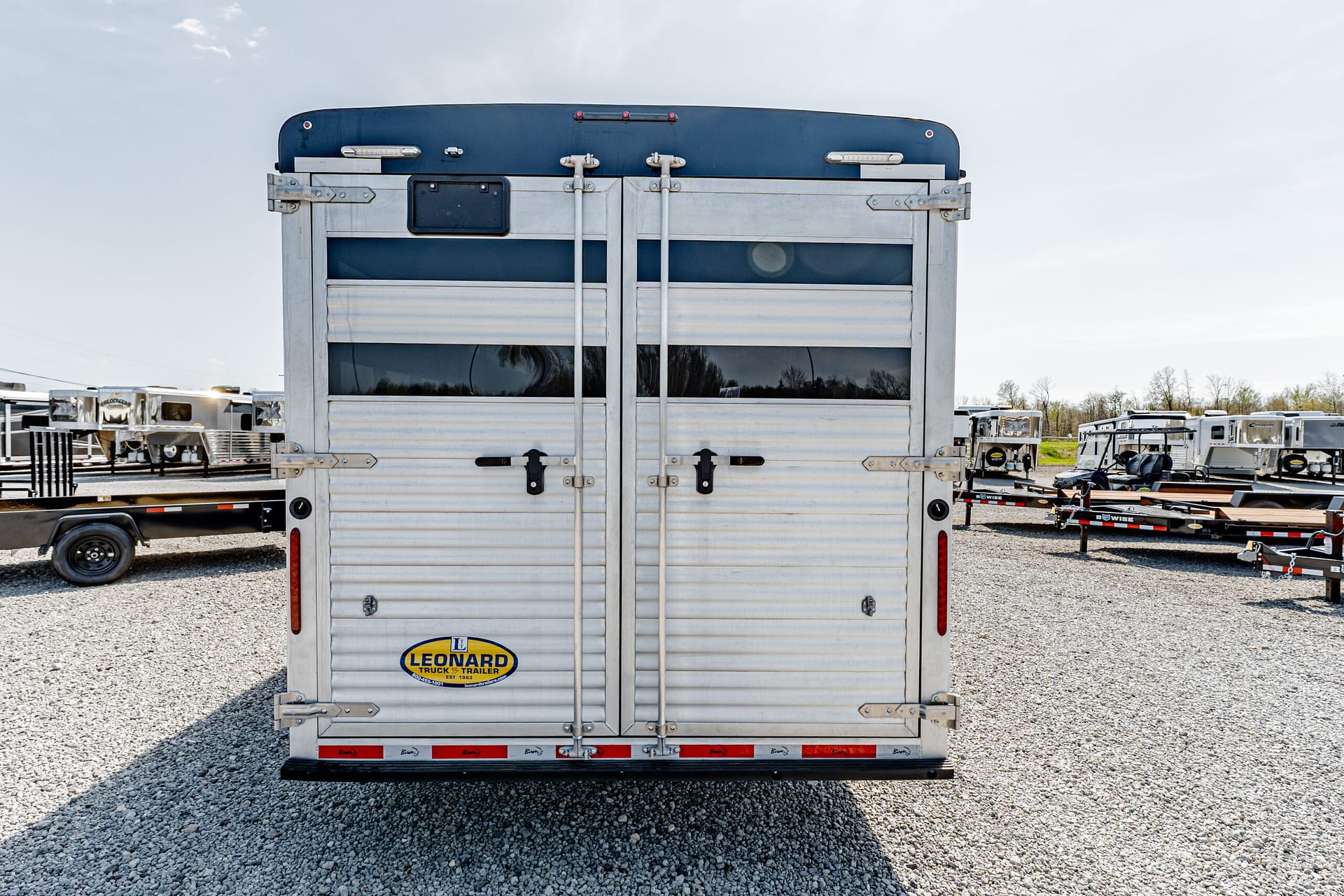 Rear view of a silver horse trailer with a blue top and Leonard logo in a gravel trailer lot.