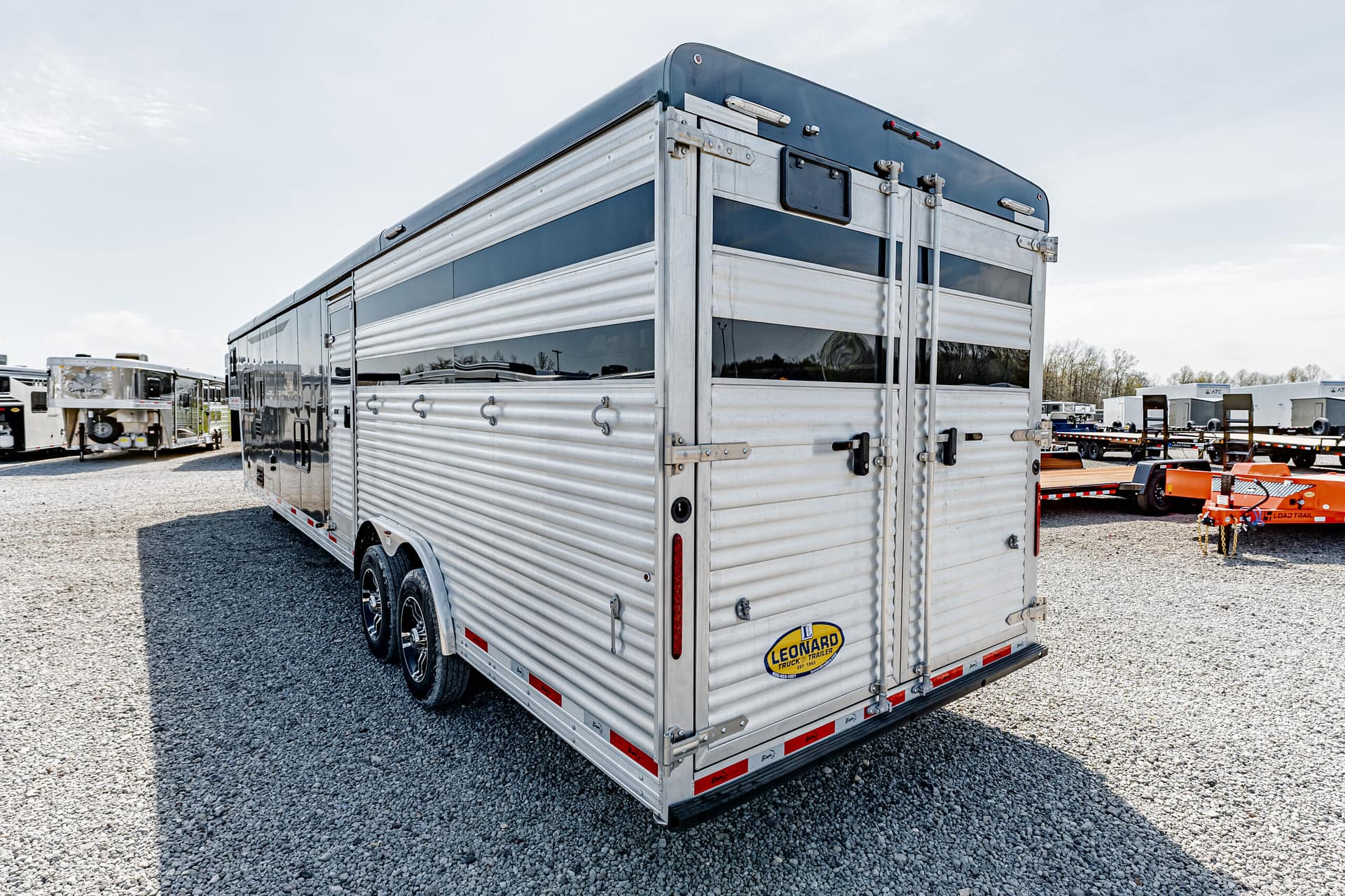 Rear of a large silver aluminum trailer with dual doors and a Leonard logo, in a gravel trailer yard with other trailers in the background.