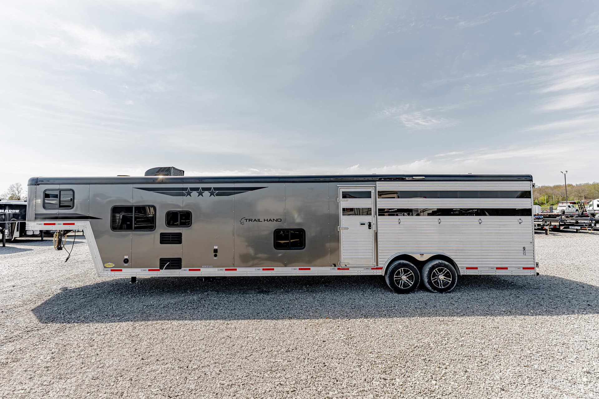 Long silver horse trailer parked on a gravel lot with a clear blue sky; other trailers visible in the background.