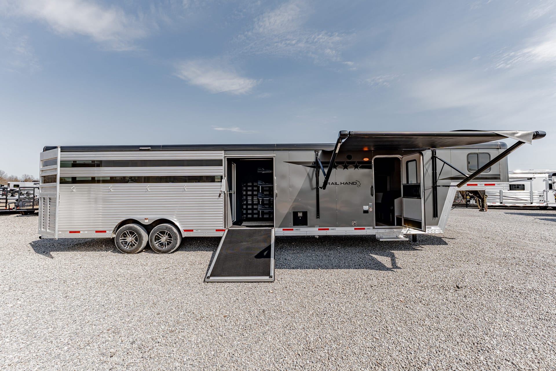 Silver horse trailer with the ramp down on a gravel lot beneath a clear blue sky