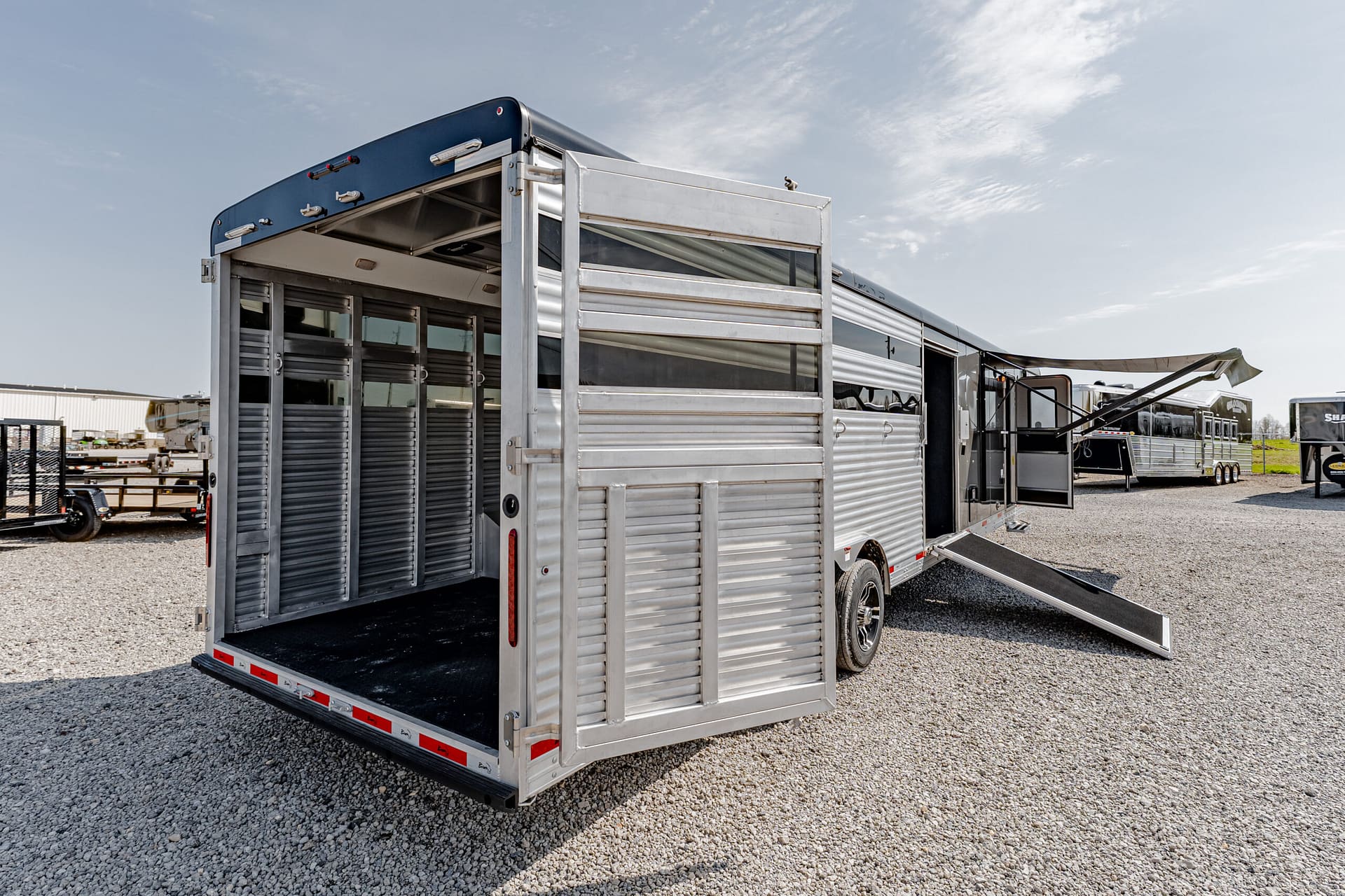 Silver aluminum livestock trailer with its rear ramp down, parked on a gravel lot under a clear sky, ready for loading or unloading.