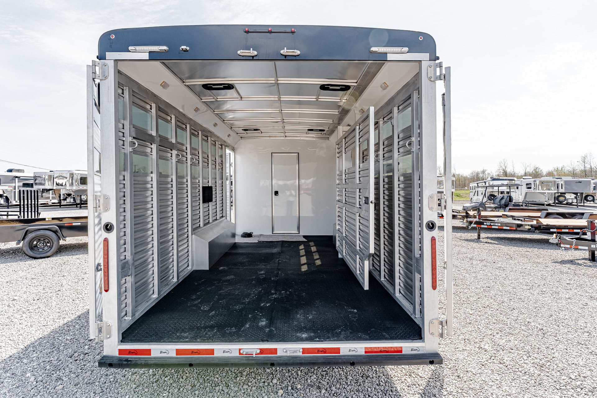 Open rear of a horse trailer showing side stalls with metal bars and a central aisle, outdoors in a trailer yard.