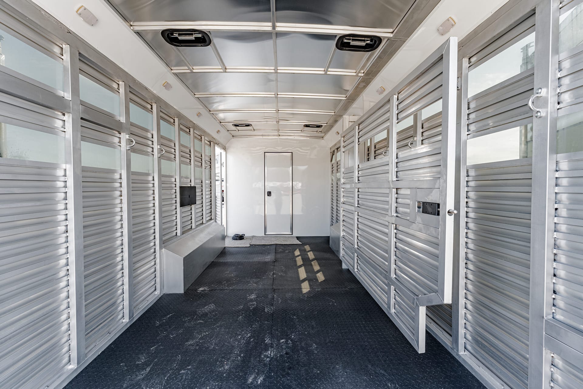 Interior of a metal horse trailer with stalls along both sides and a central aisle toward a back door