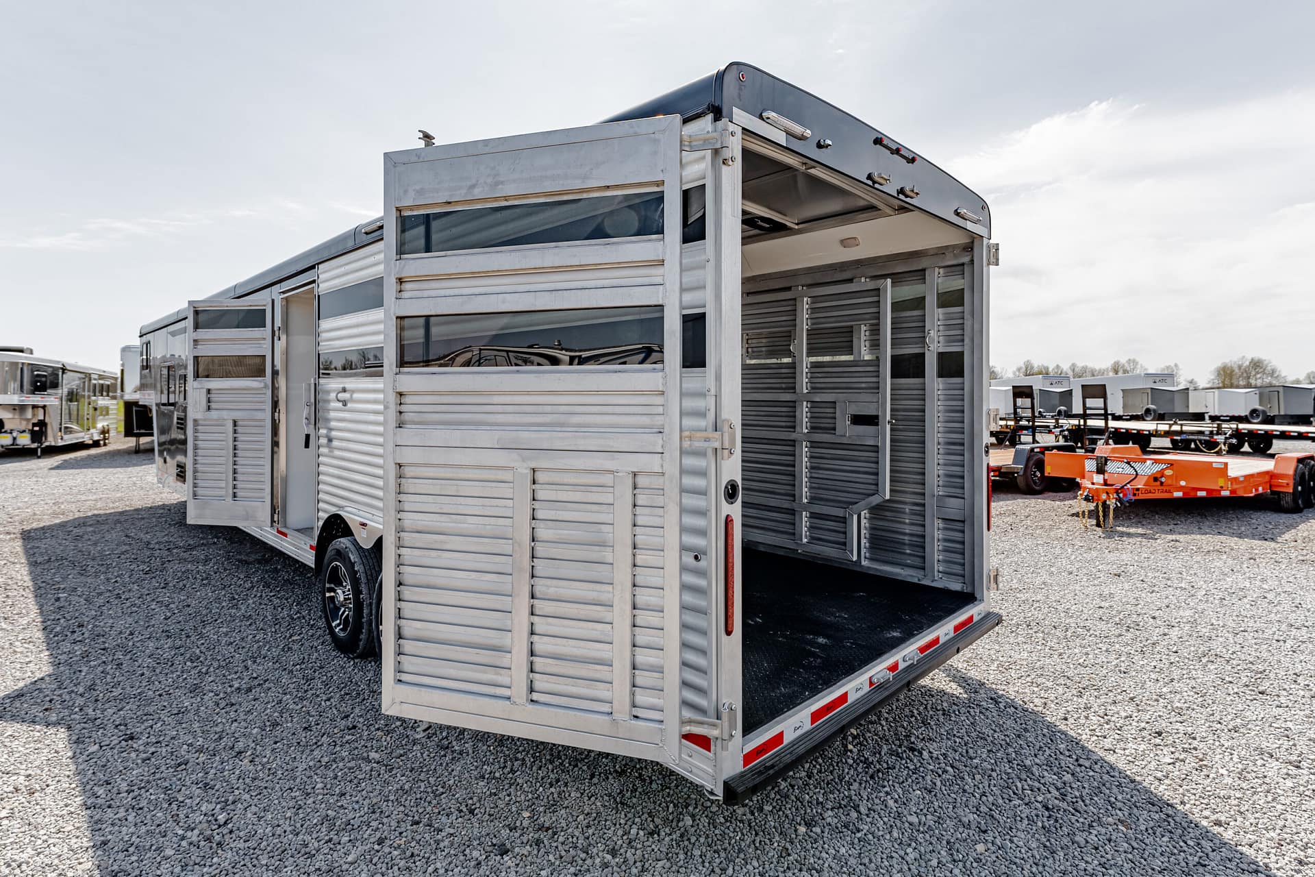 Rear view of a silver aluminum livestock trailer with double doors open, revealing interior stalls on a gravel lot.