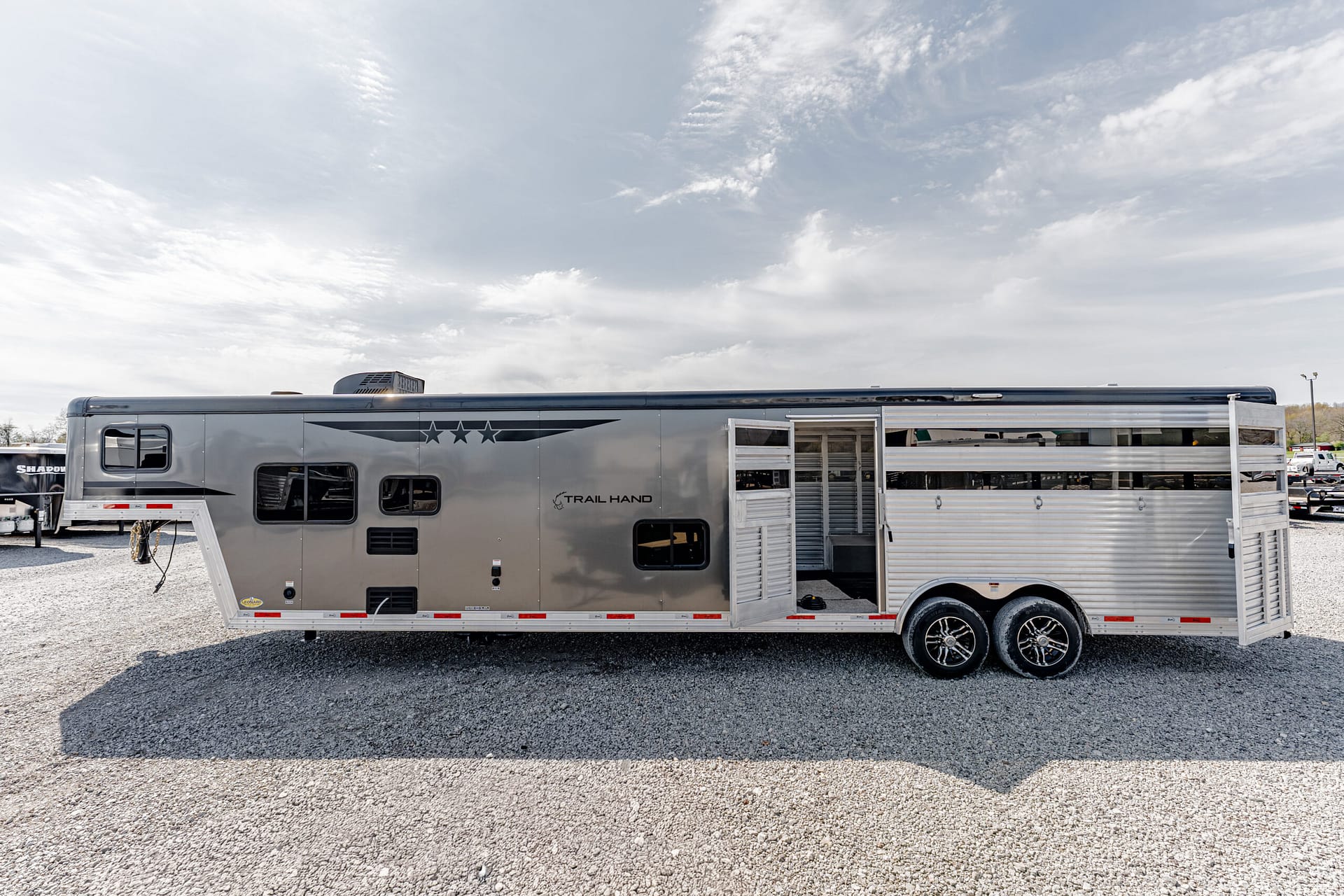 Silver travel trailer with extended side slide-out parked on a gravel lot under a cloudy sky.