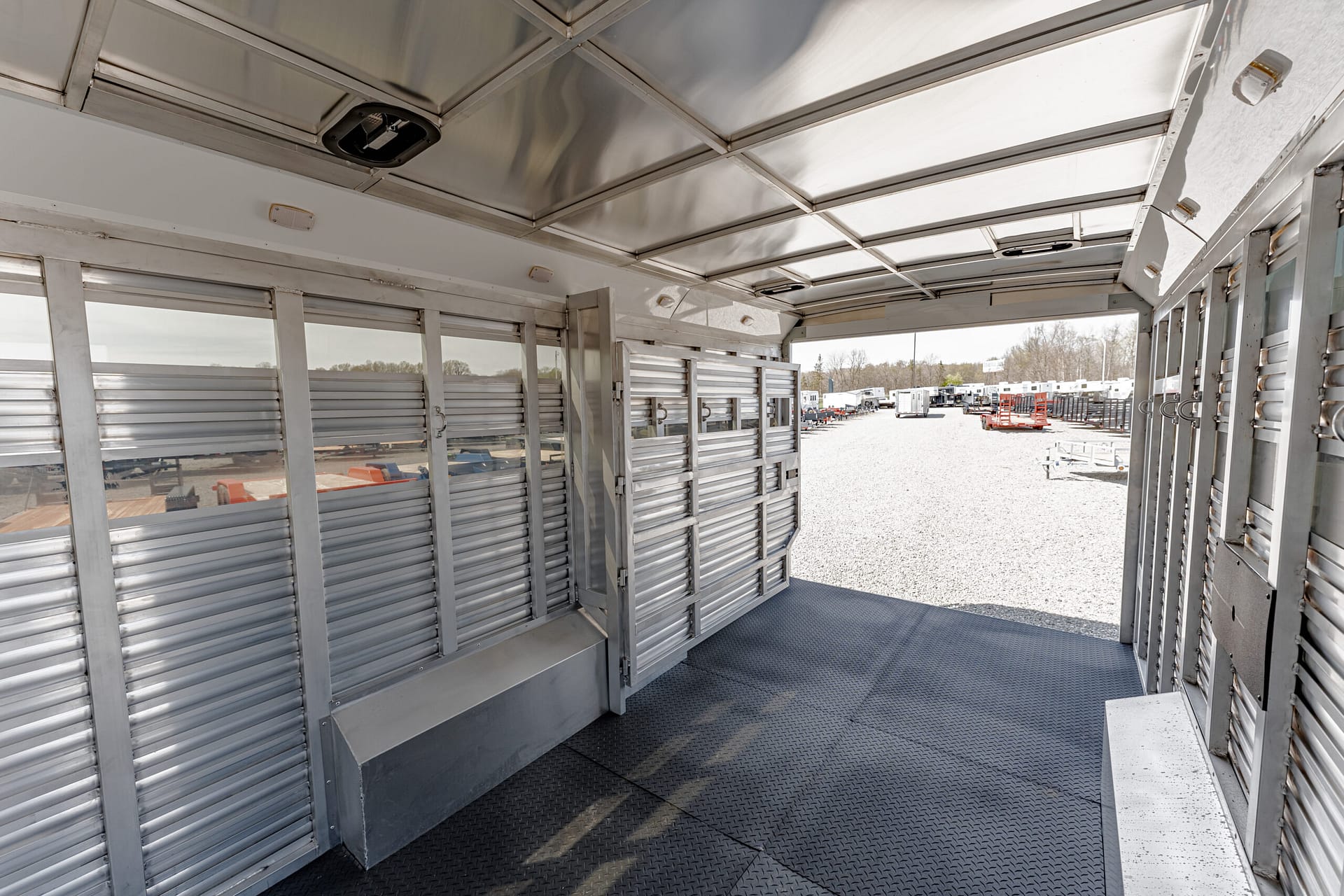 Interior of a metallic livestock trailer with ribbed aluminum walls; open rear doorway shows a gravel yard with parked trailers outside.