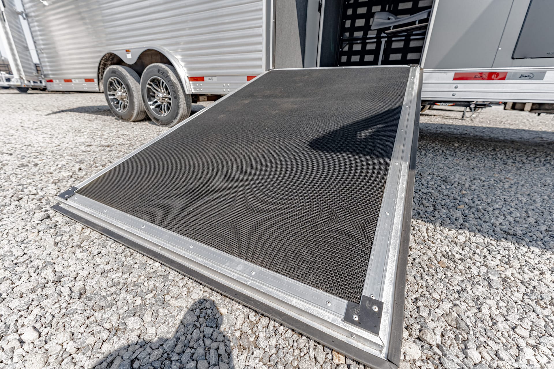 Metal loading ramp extending from a silver trailer onto a gravel lot, with another trailer visible in the background.