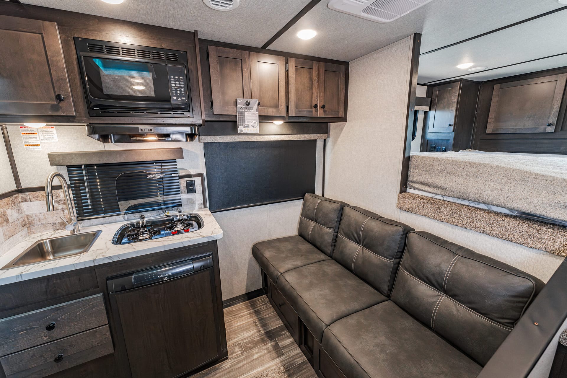 Compact RV kitchen and living area: microwave, stovetop, sink, dark wood cabinets, and a dark leather sofa with a bed area in the background.