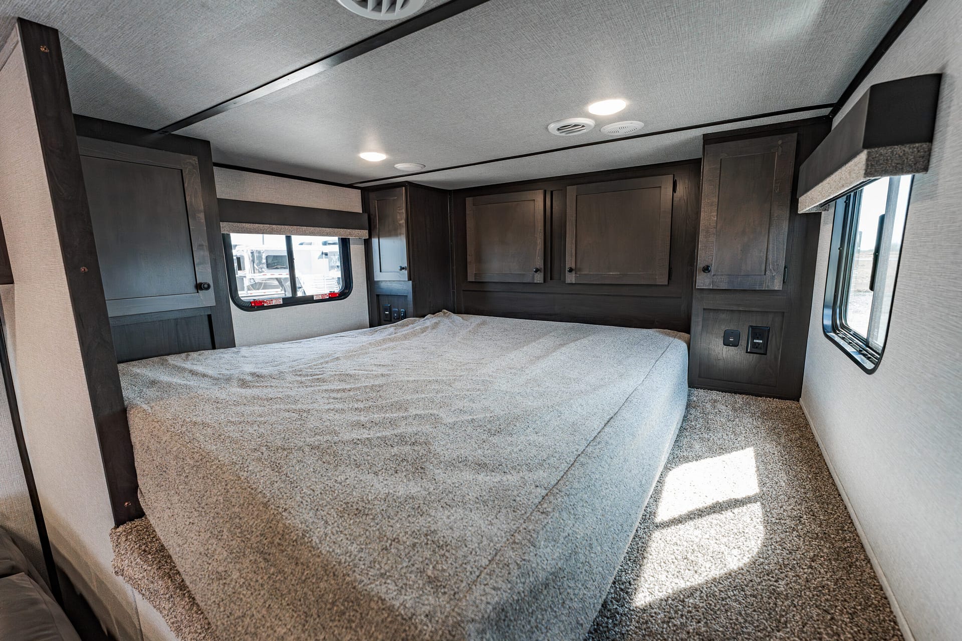 Interior of a small RV bedroom with a carpeted bed and dark wood cabinets along the wall, window on the left, ceiling lights above